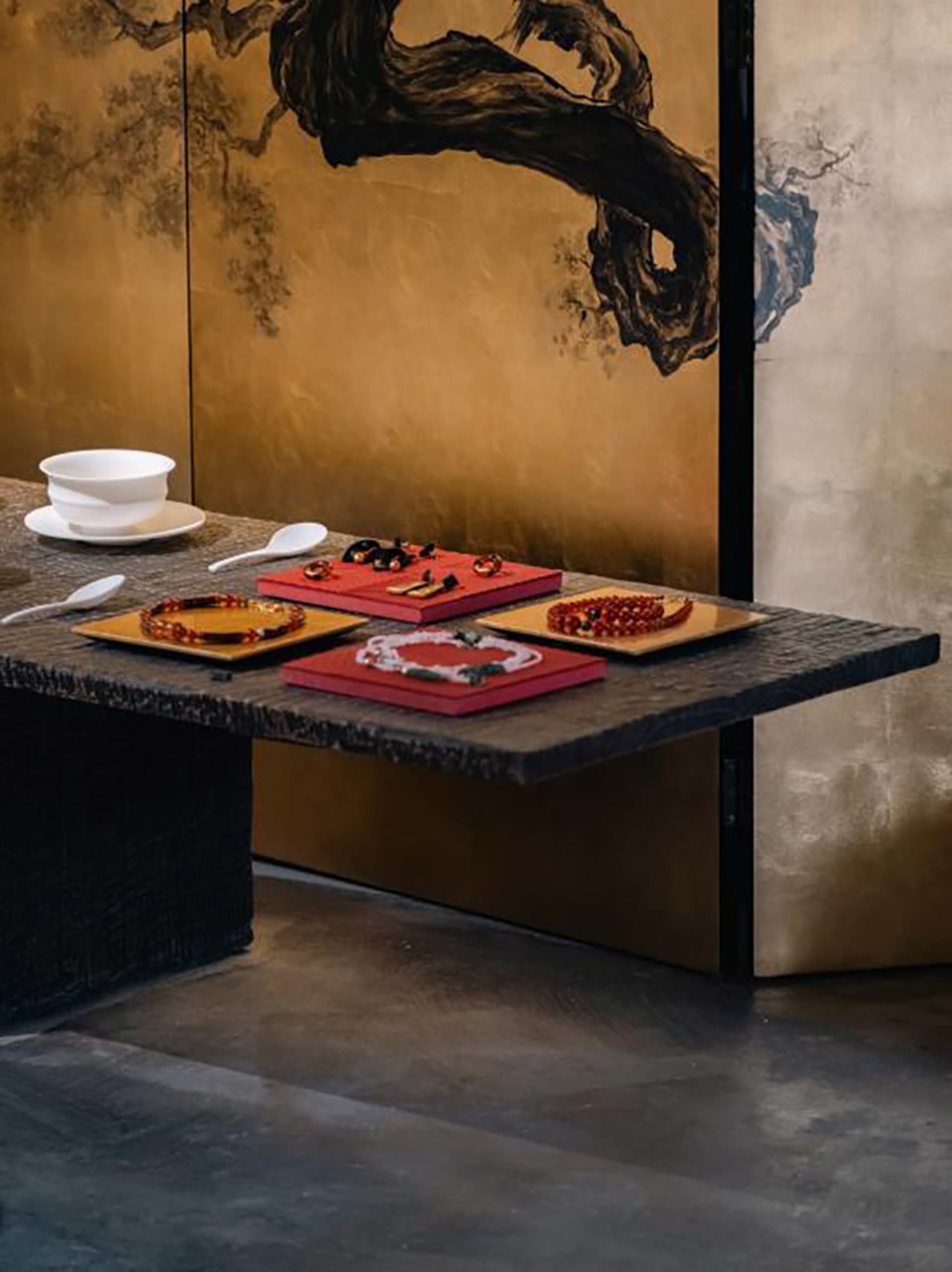 Dark stone console table displaying colorful jewelry on red and gold trays, set against a luxurious gold-leaf folding screen with traditional ink-wash landscape painting.