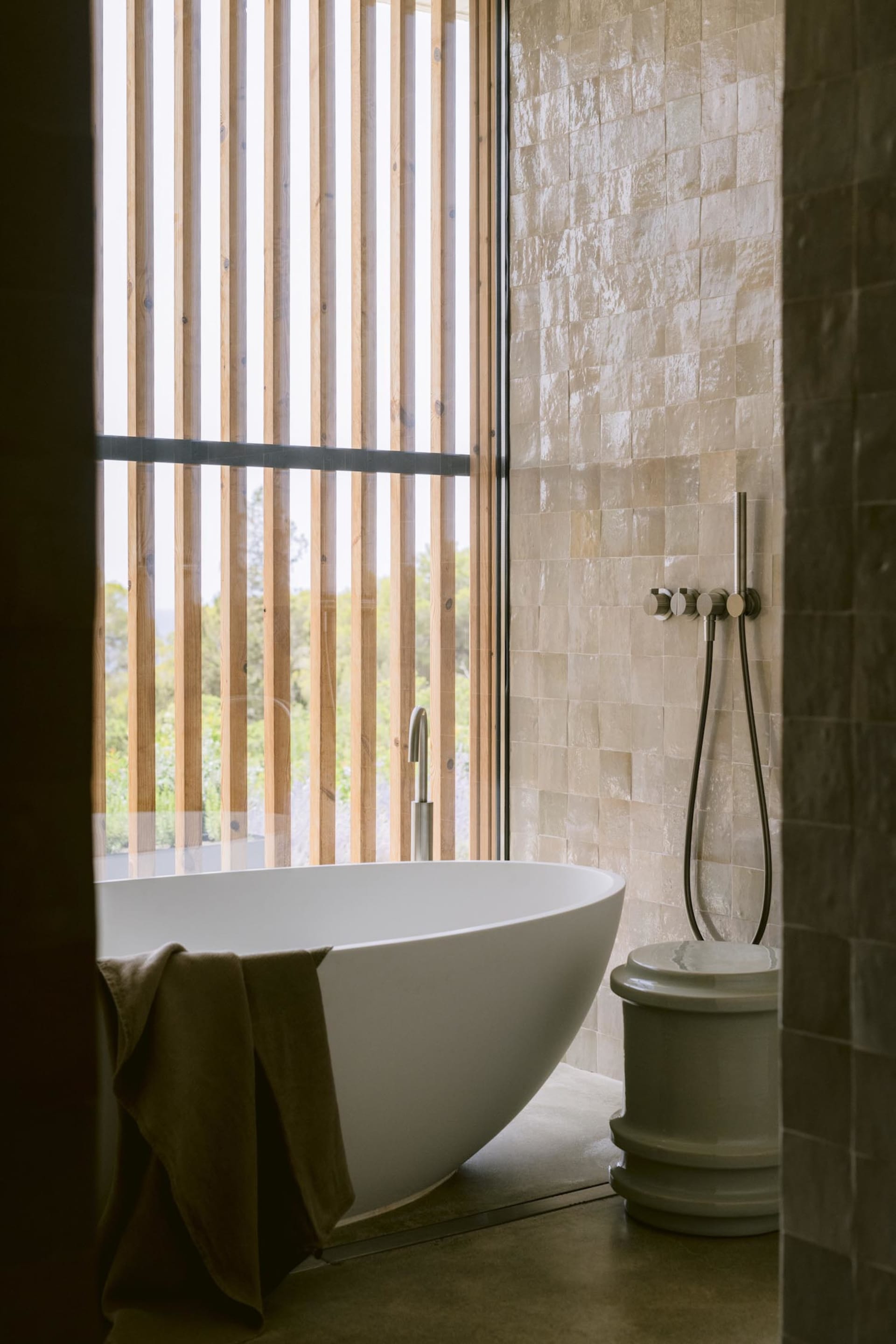 Modern minimalist bathroom featuring a white freestanding oval bathtub, beige travertine tiled walls, vertical wooden slats with mountain view, and a matte grey vessel toilet.