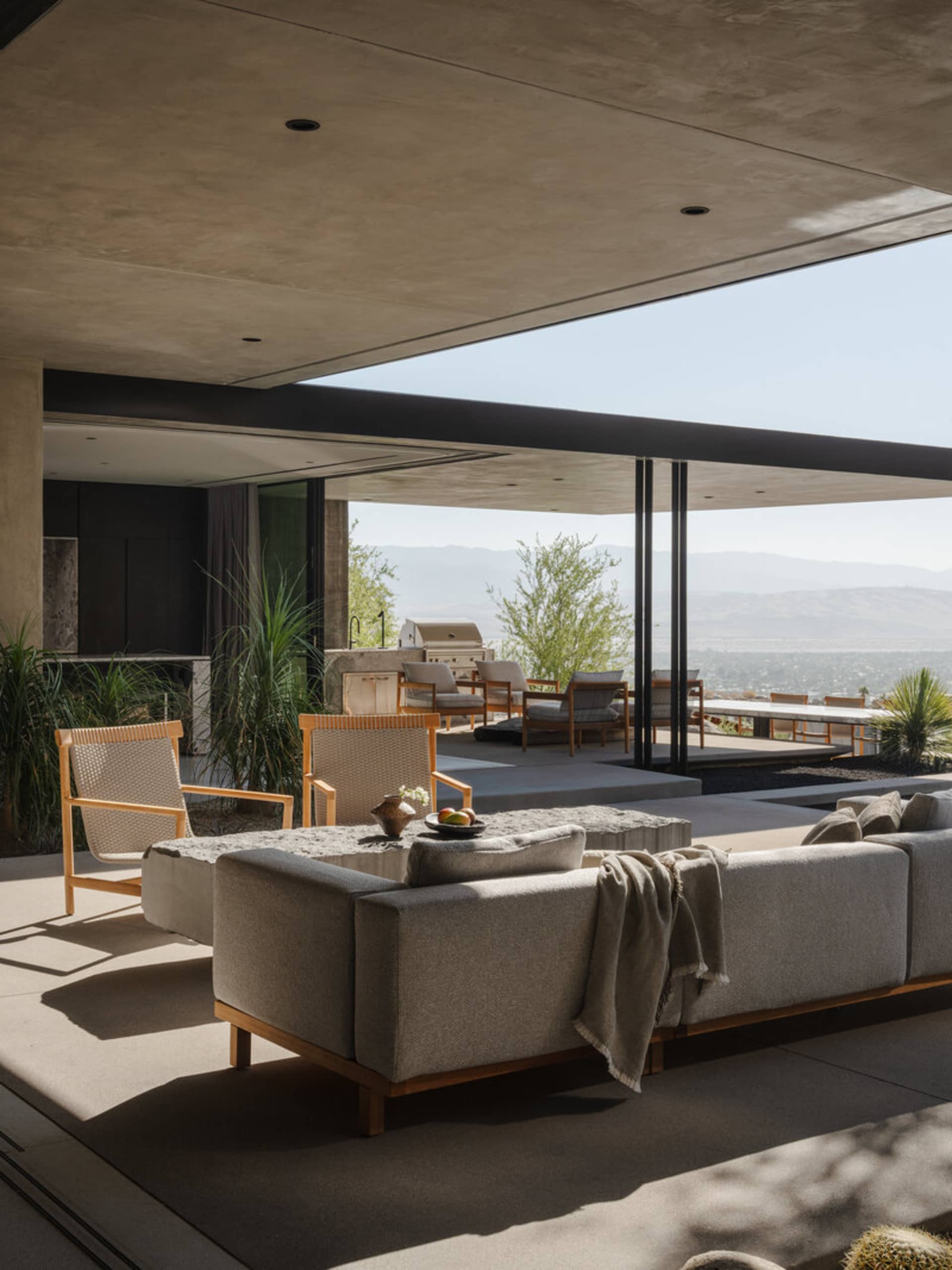 Modern outdoor living area with concrete ceiling, wooden-framed sofas, woven lounge chairs, and panoramic desert mountain views.