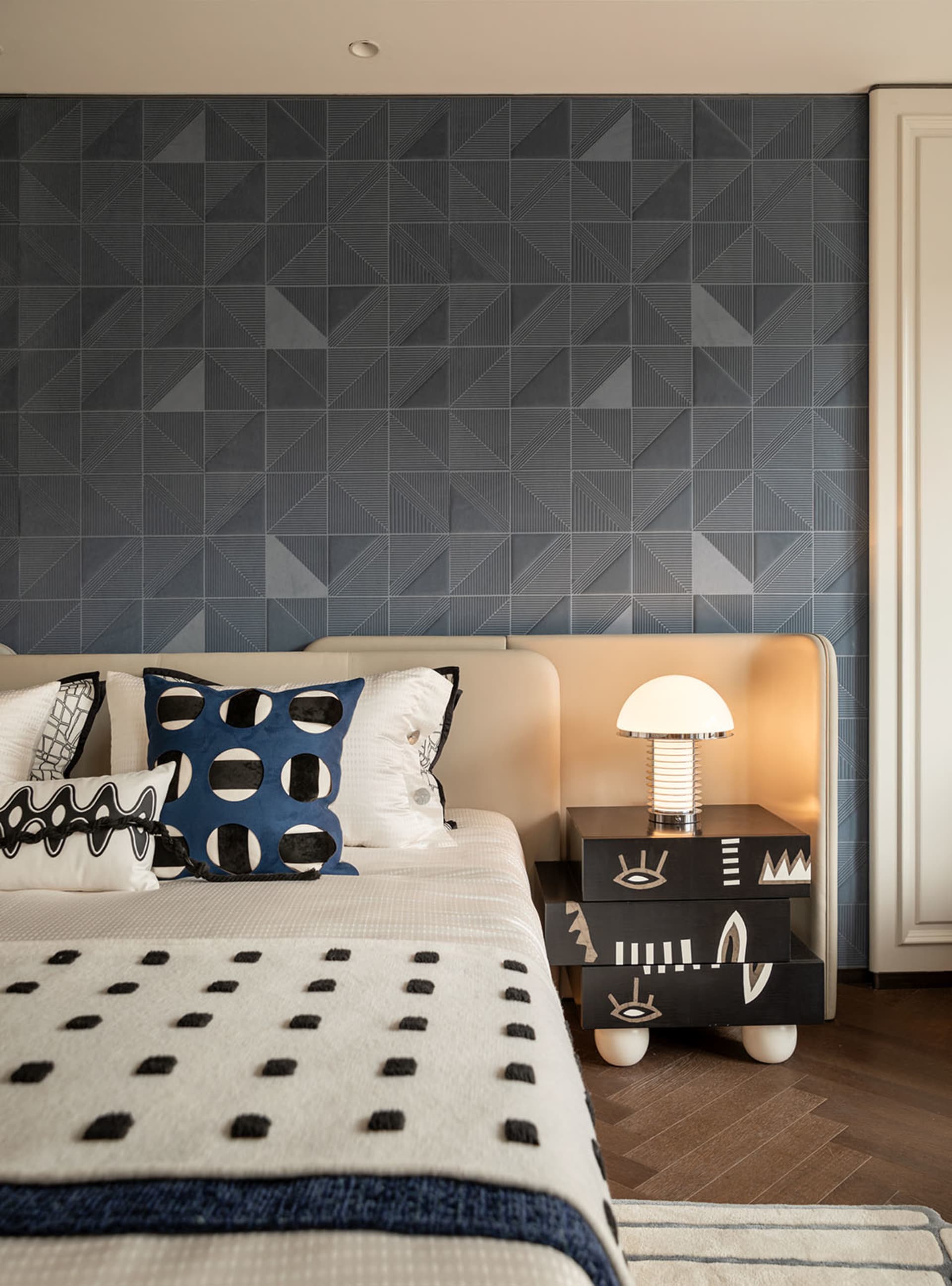Modern bedroom interior with dark navy geometric patterned tile accent wall, beige upholstered bed with blue and black patterned pillows, and a black graphic nightstand with a modern mushroom lamp.