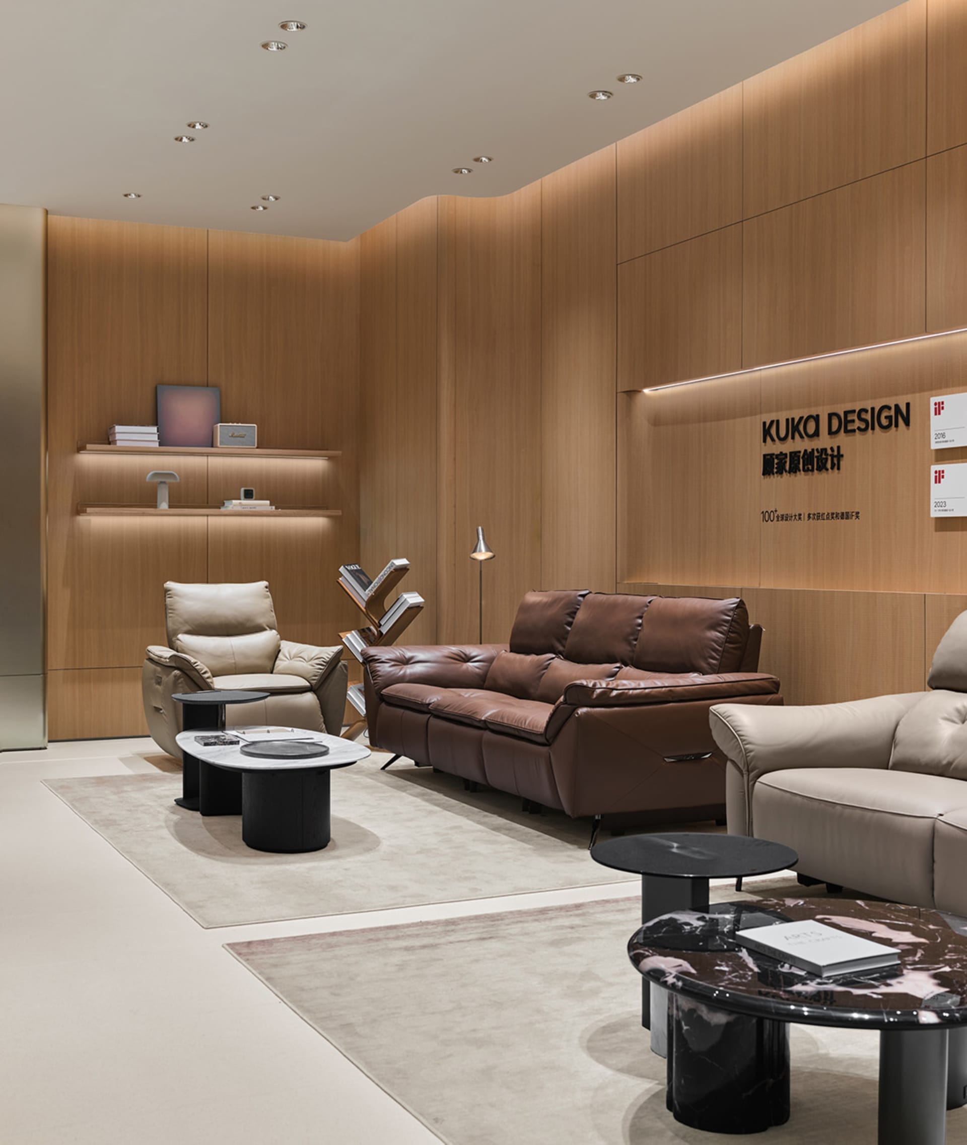 Modern luxury living room display with brown leather sofa, cream leather armchairs, black marble coffee tables, and warm wood panel walls with integrated lighting.