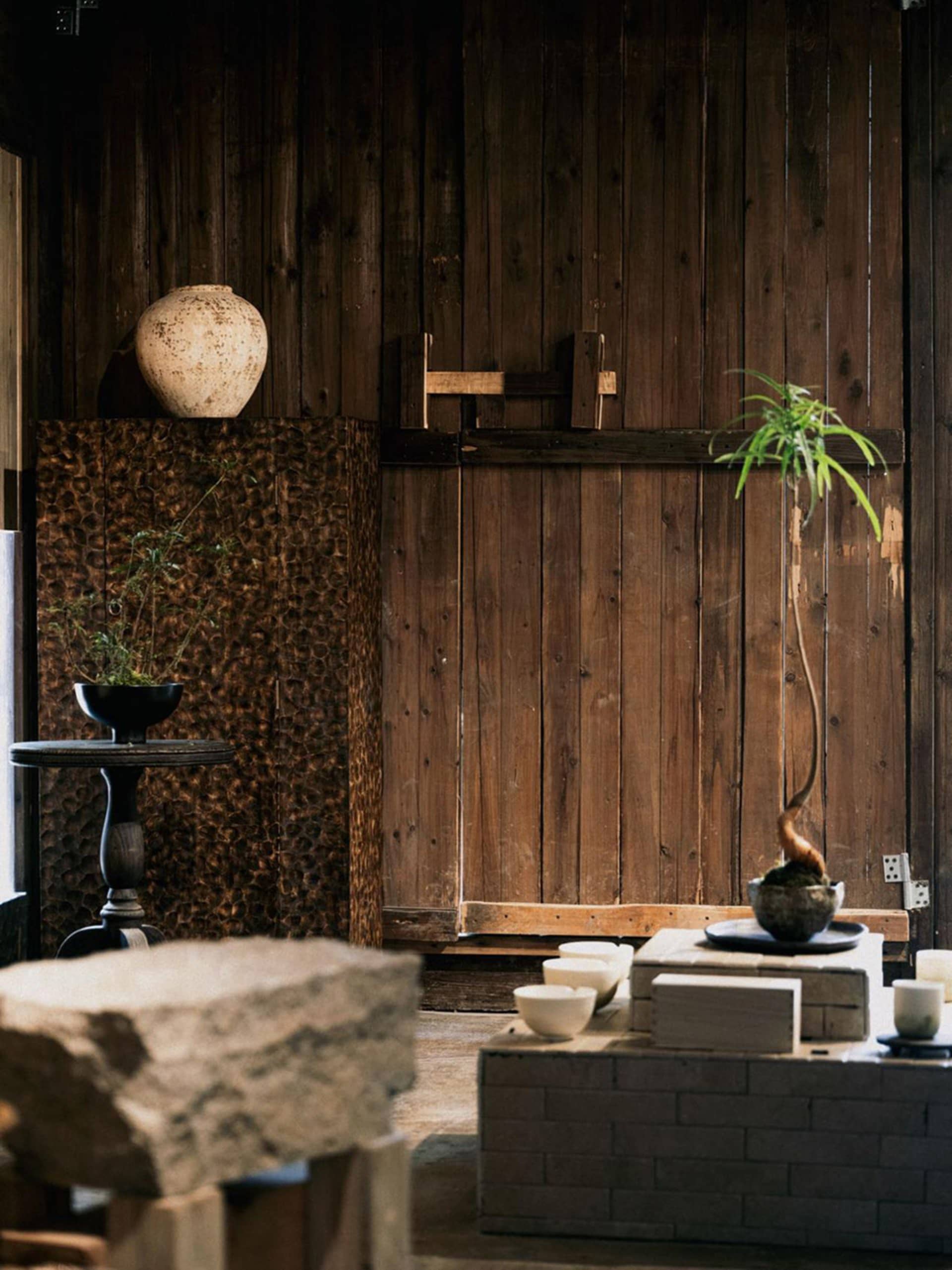 Interior of a Japanese wabi-sabi style tea room featuring dark reclaimed wood walls, a textured stone wall panel, large rough-hewn stone blocks, ceramic tea bowls on a brick platform, and potted plants in rustic vessels.