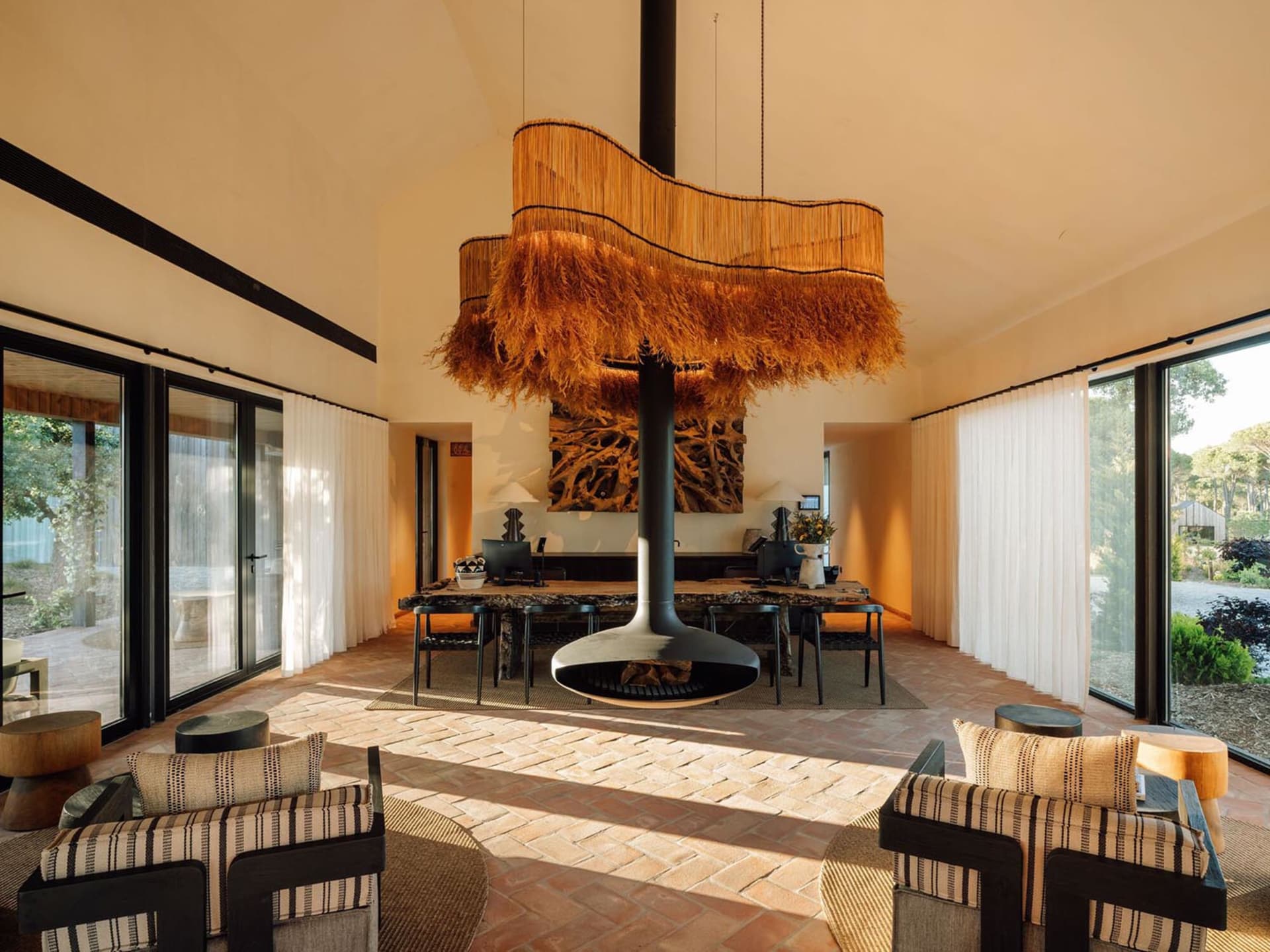 Spacious living room featuring a large woven pendant light, central black fireplace, terracotta tile floor, and striped armchairs with natural light from large glass doors.