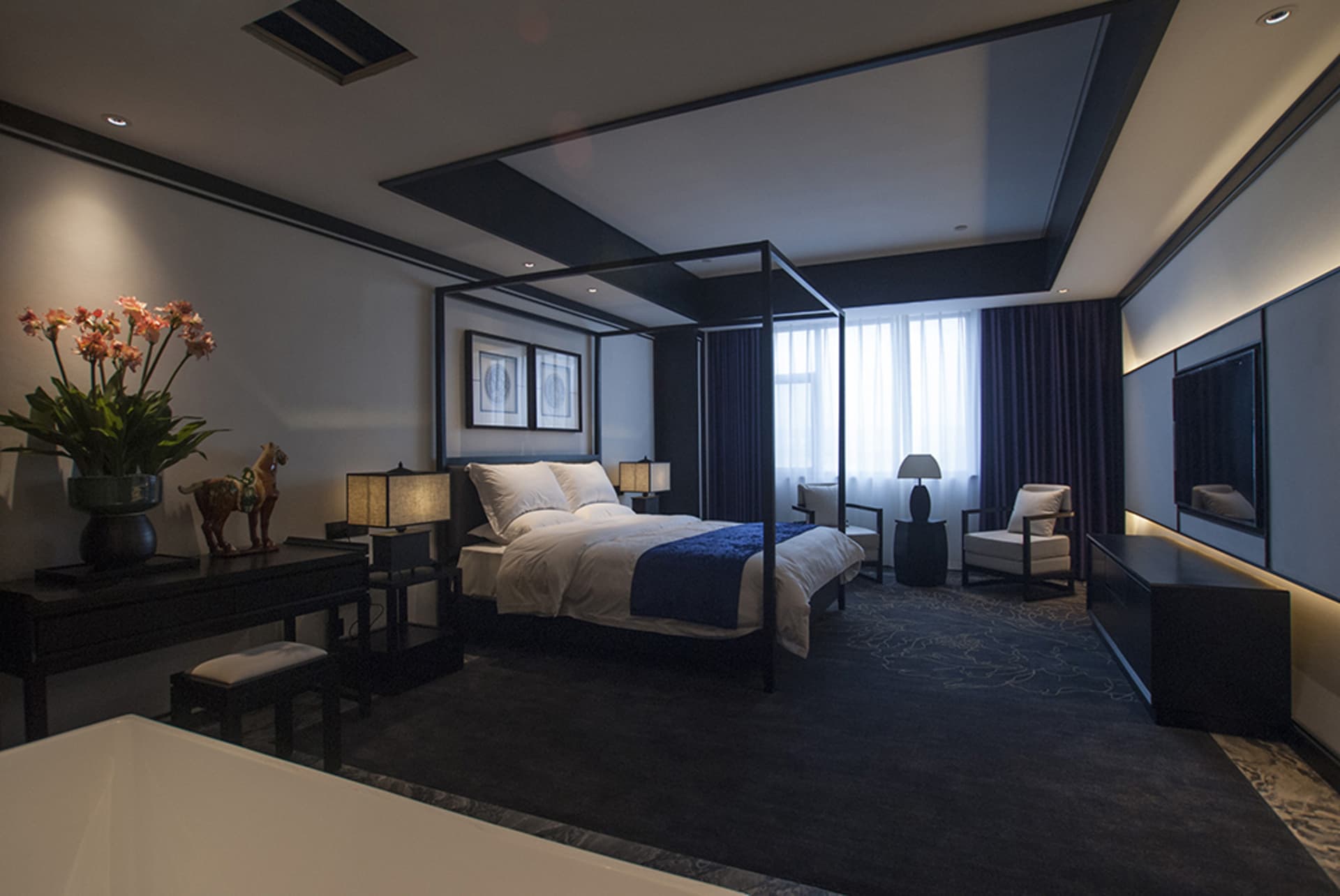 Modern luxury bedroom with black four-poster bed, dark wood furniture, white and navy bedding, and blue patterned carpet.