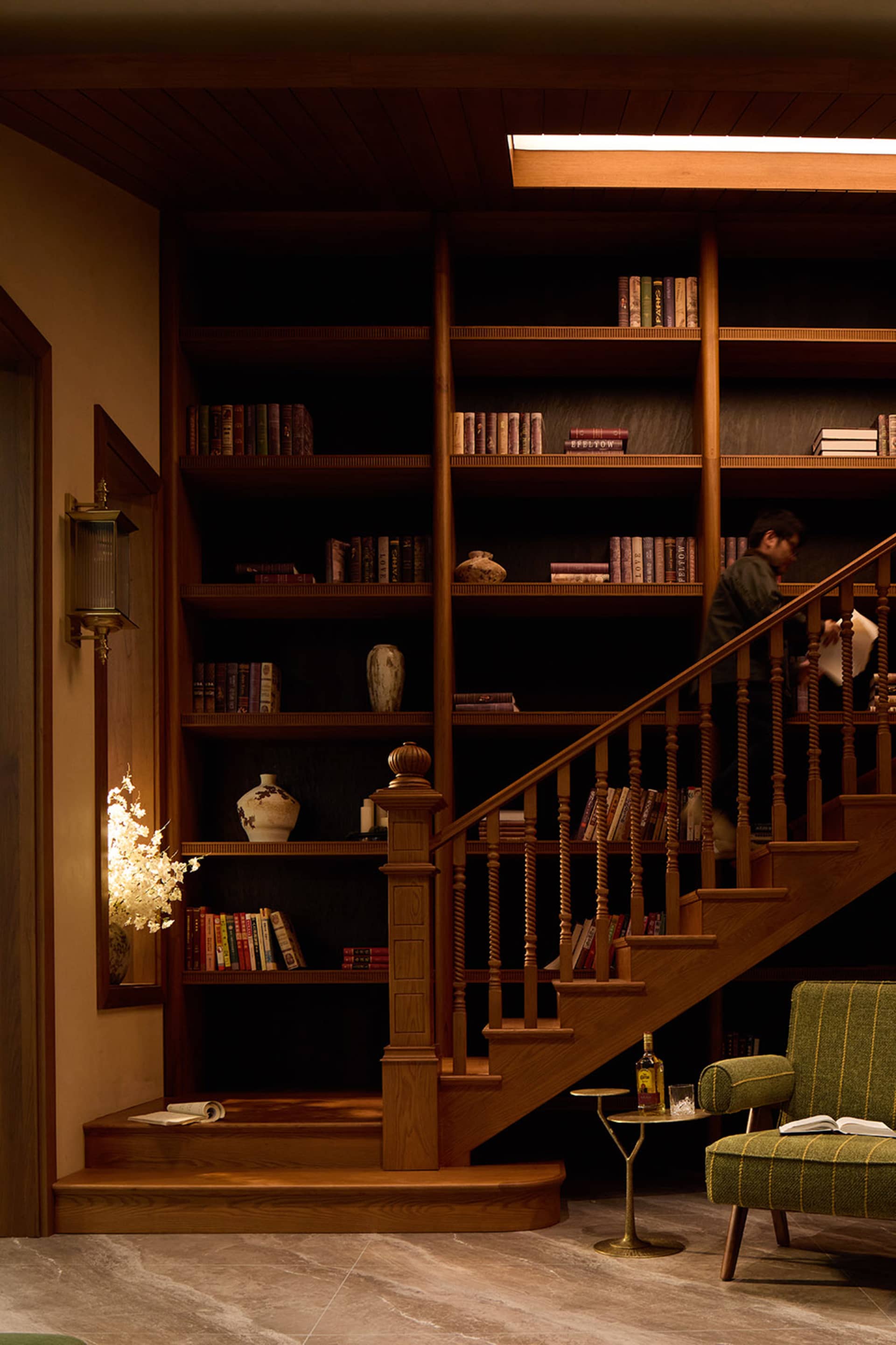 Warmly lit interior featuring a walnut staircase with integrated bookshelves, a green striped armchair, and a person browsing books on the upper level.