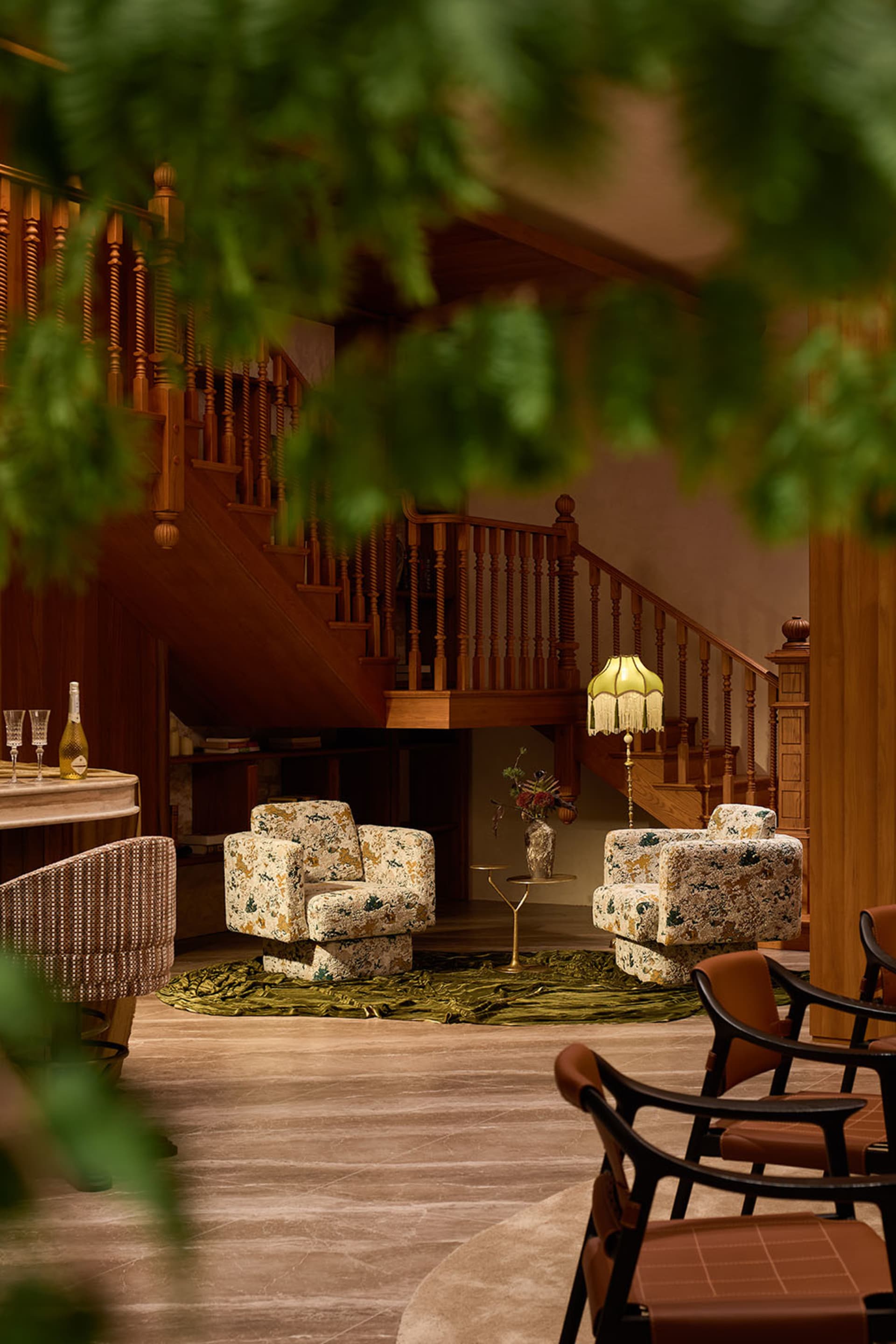 Interior view of a luxurious lounge area featuring two floral-patterned armchairs, a green velvet rug, wooden staircase with turned balusters, and warm ambient lighting from a fringed floor lamp.