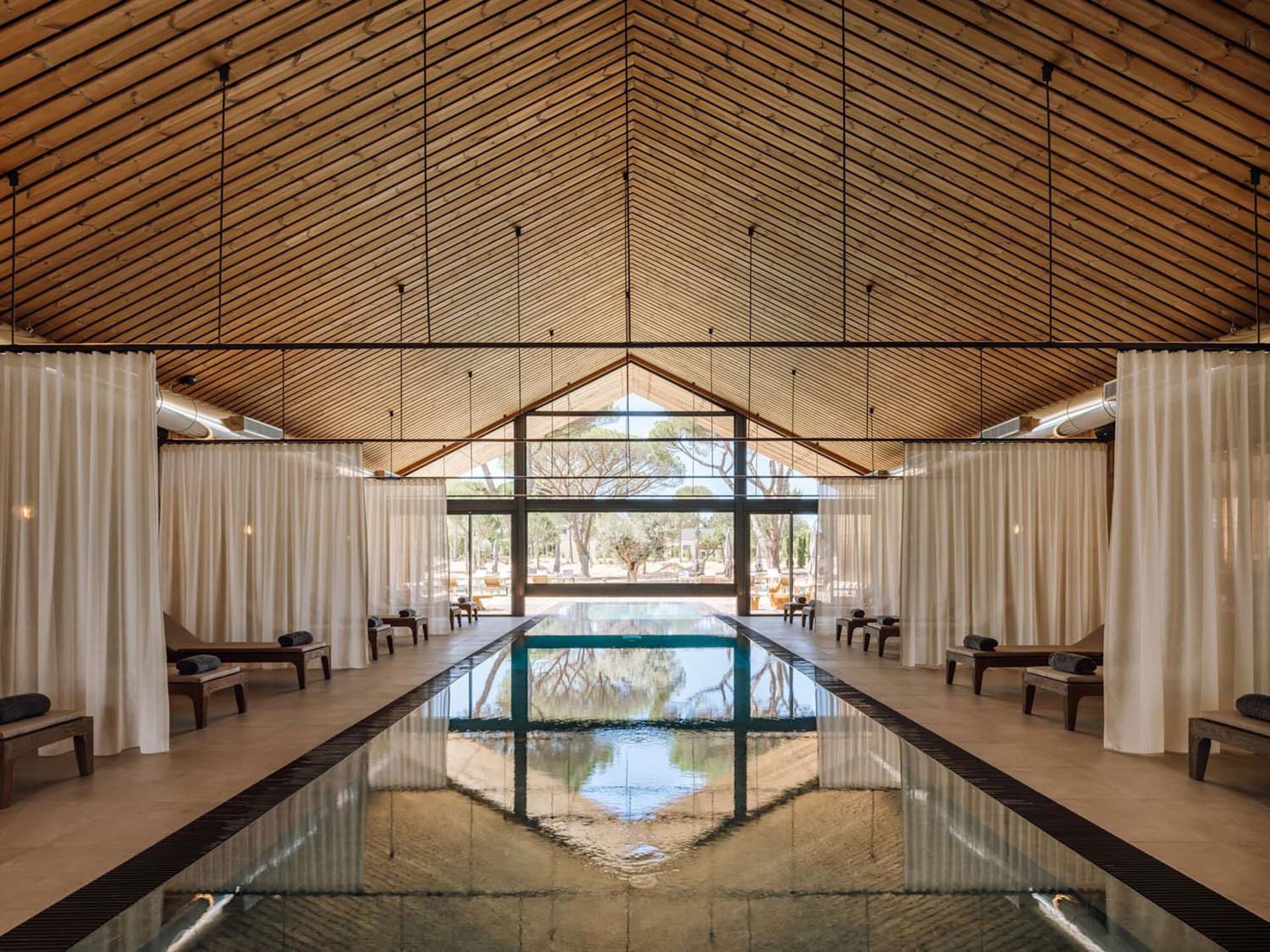 Long indoor lap pool in a serene spa with high vaulted wooden ceiling, white curtains, and wooden lounge chairs on both sides.