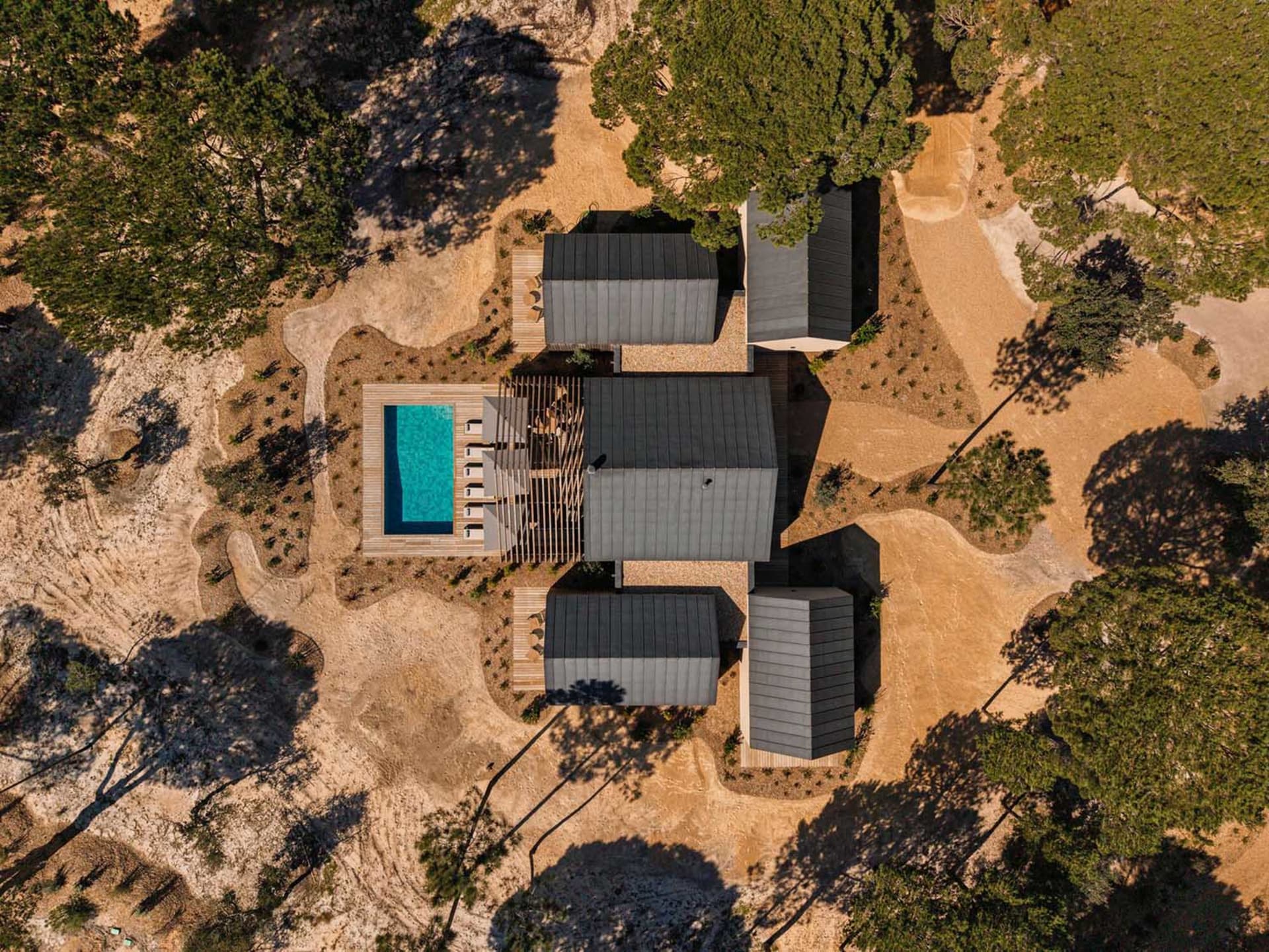 Aerial view of a modern desert villa featuring dark metal roofs, a rectangular turquoise swimming pool, wooden decks, and surrounded by arid landscape with scattered pine trees.