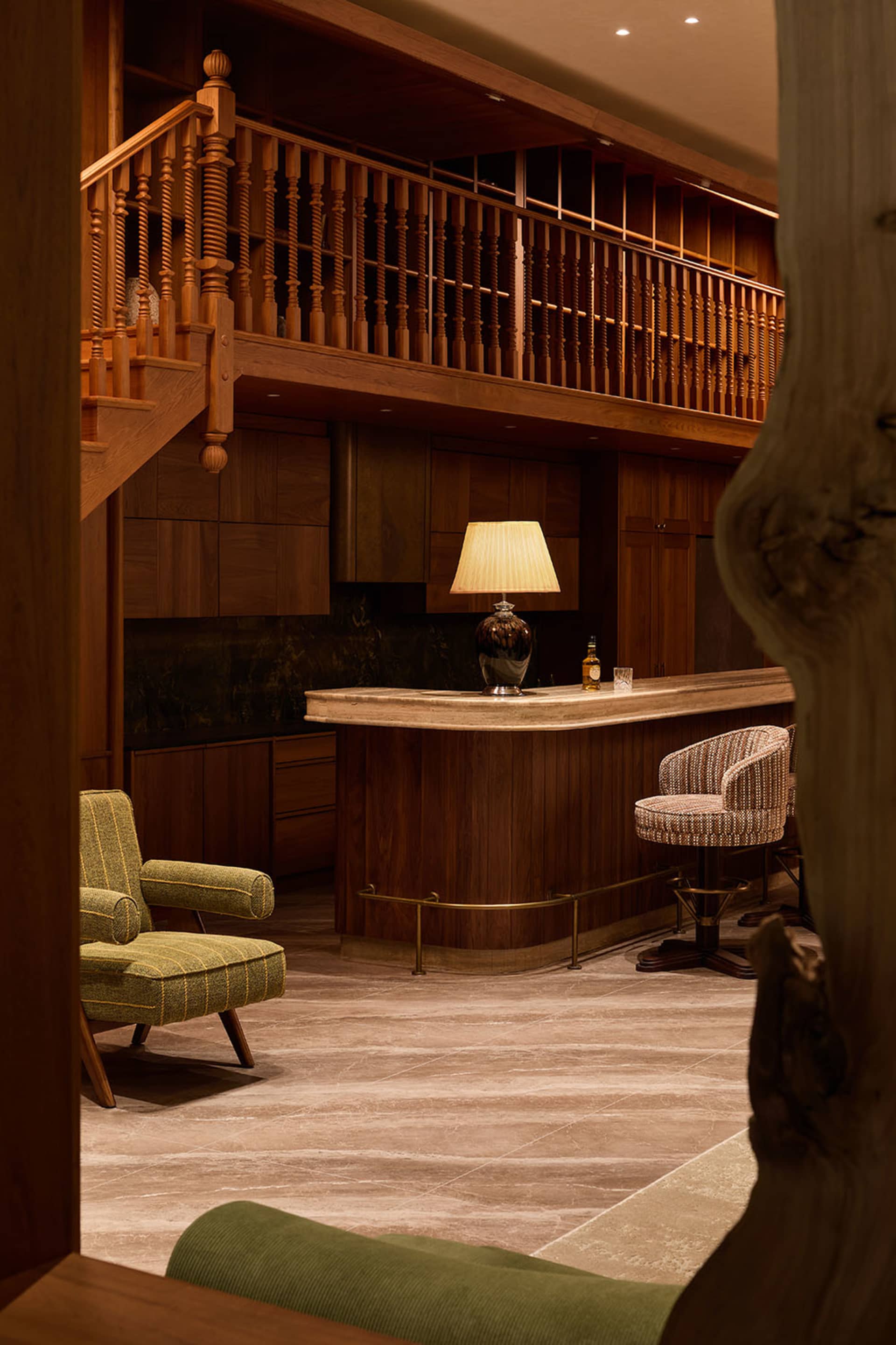 Luxurious residential home bar with rich walnut wood paneling, a curved bar counter, green striped armchair, patterned swivel bar stools, and a two-story wooden mezzanine railing.