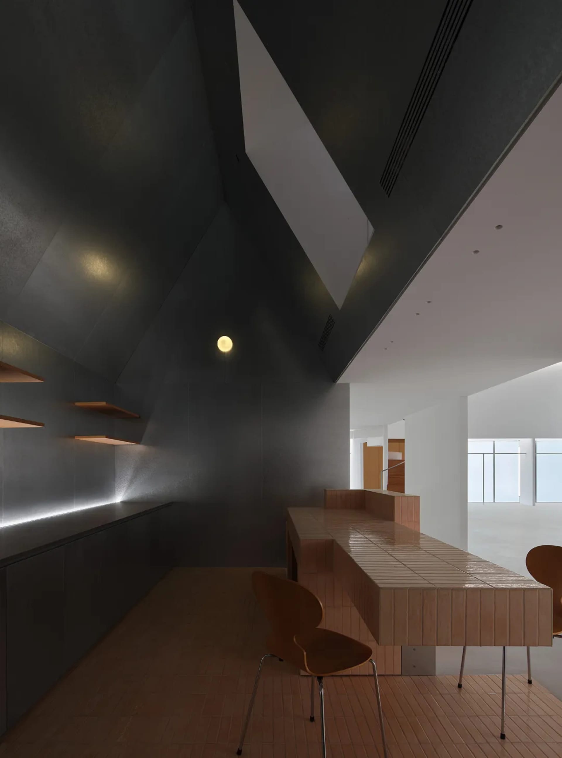 Modern minimalist dining room featuring dark concrete walls, a long light-wood tiled dining table with built-in banquette, wooden chairs, and dramatic angular architectural lines.
