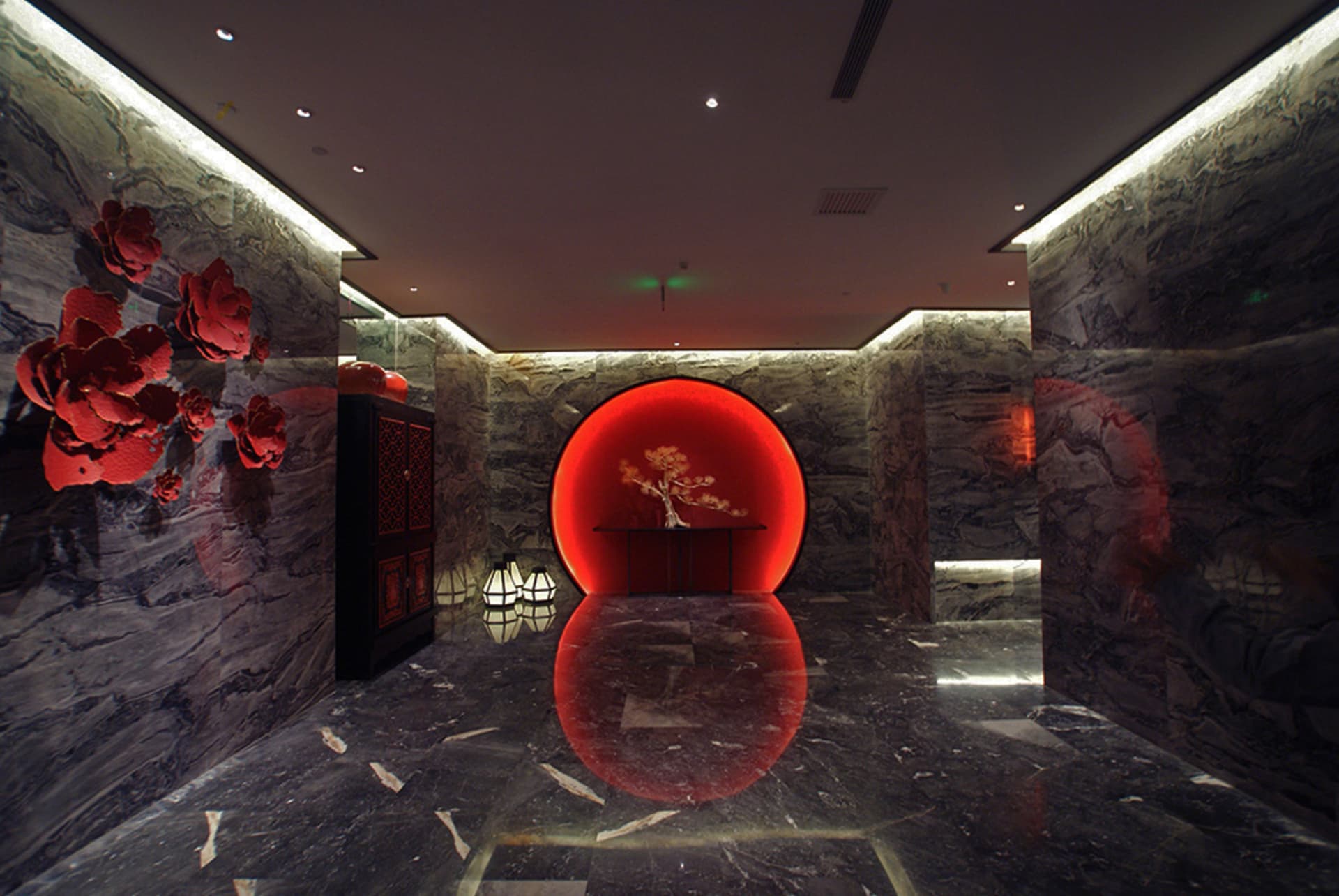 Luxurious modern Chinese-style lobby with dark marble walls and floor, featuring a glowing red circular moon gate with a bonsai tree inside, large red floral wall art, and black lacquered cabinet.