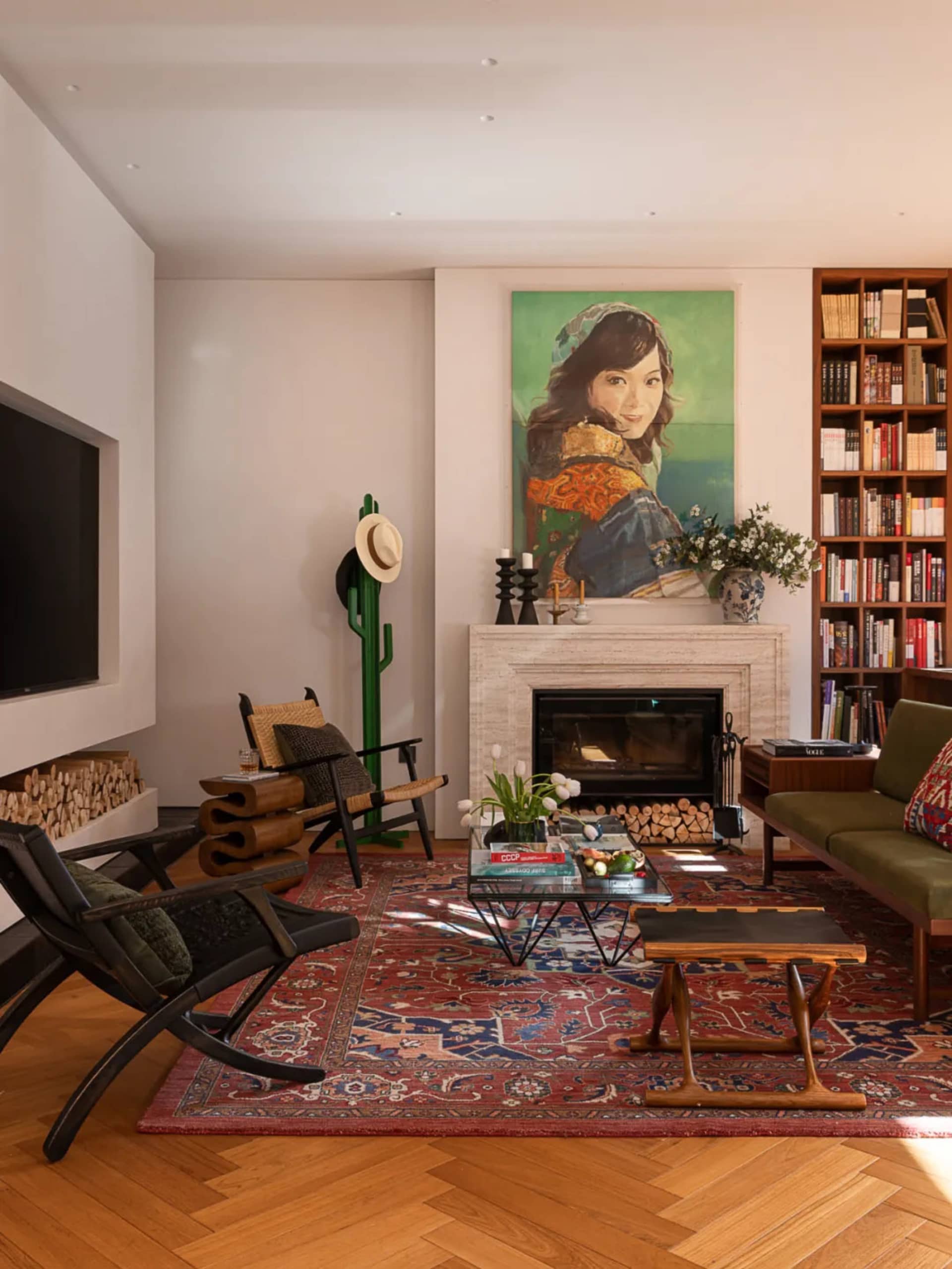 Eclectic living room featuring a large red Persian rug, marble fireplace, green velvet sofa, black leather lounge chairs, built-in bookshelves, and a colorful portrait painting above the mantel.