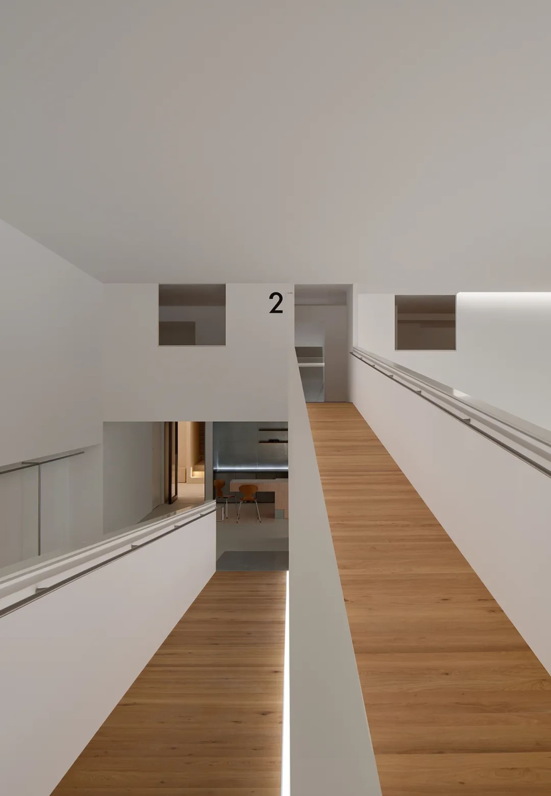 Modern double-height interior corridor featuring light oak wood flooring, white walls, continuous handrails, and linear LED lighting strips along the floor edge.