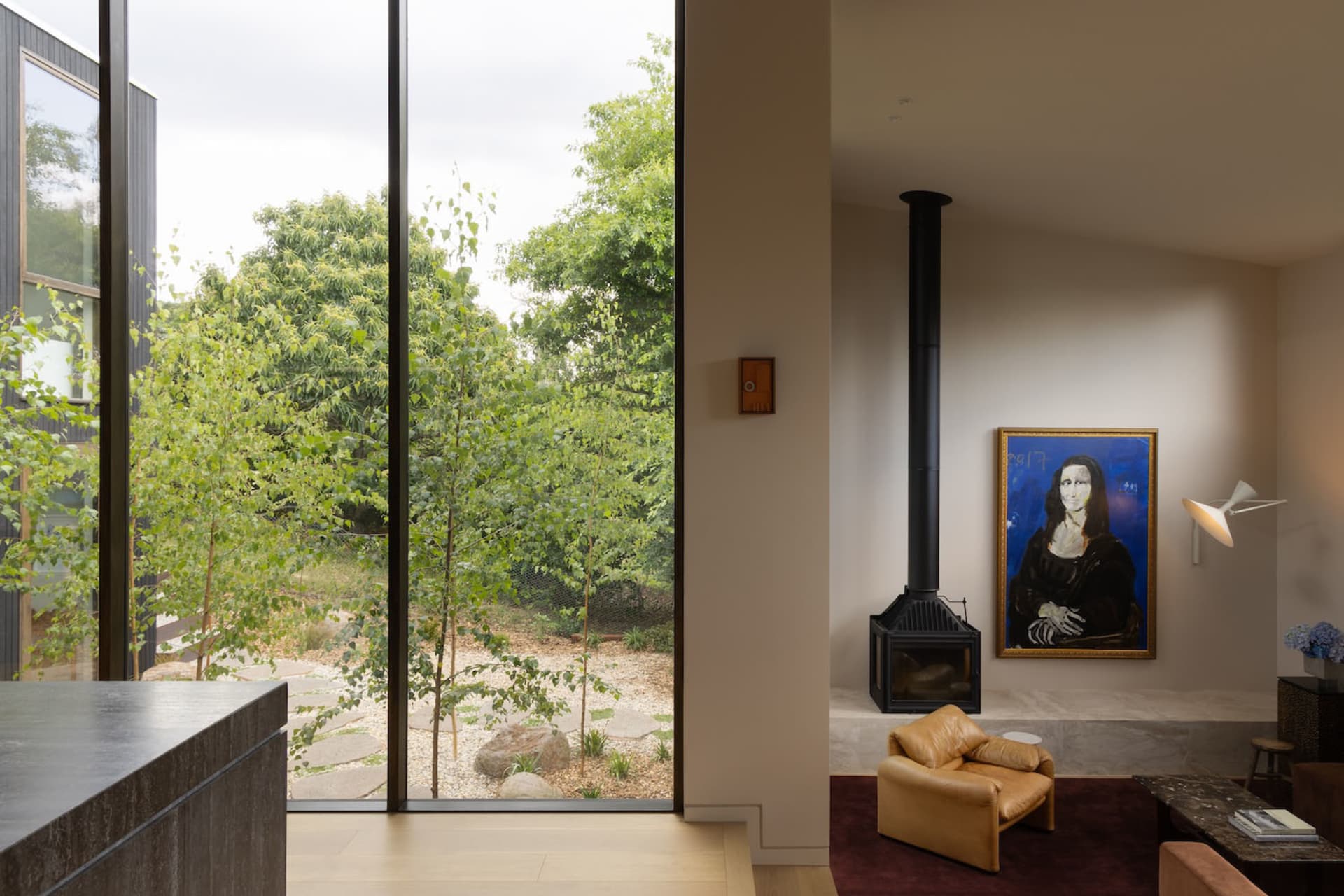 Contemporary living room featuring a tan leather armchair, black metal fireplace, large Mona Lisa painting, and floor-to-ceiling glass doors opening to a green garden.