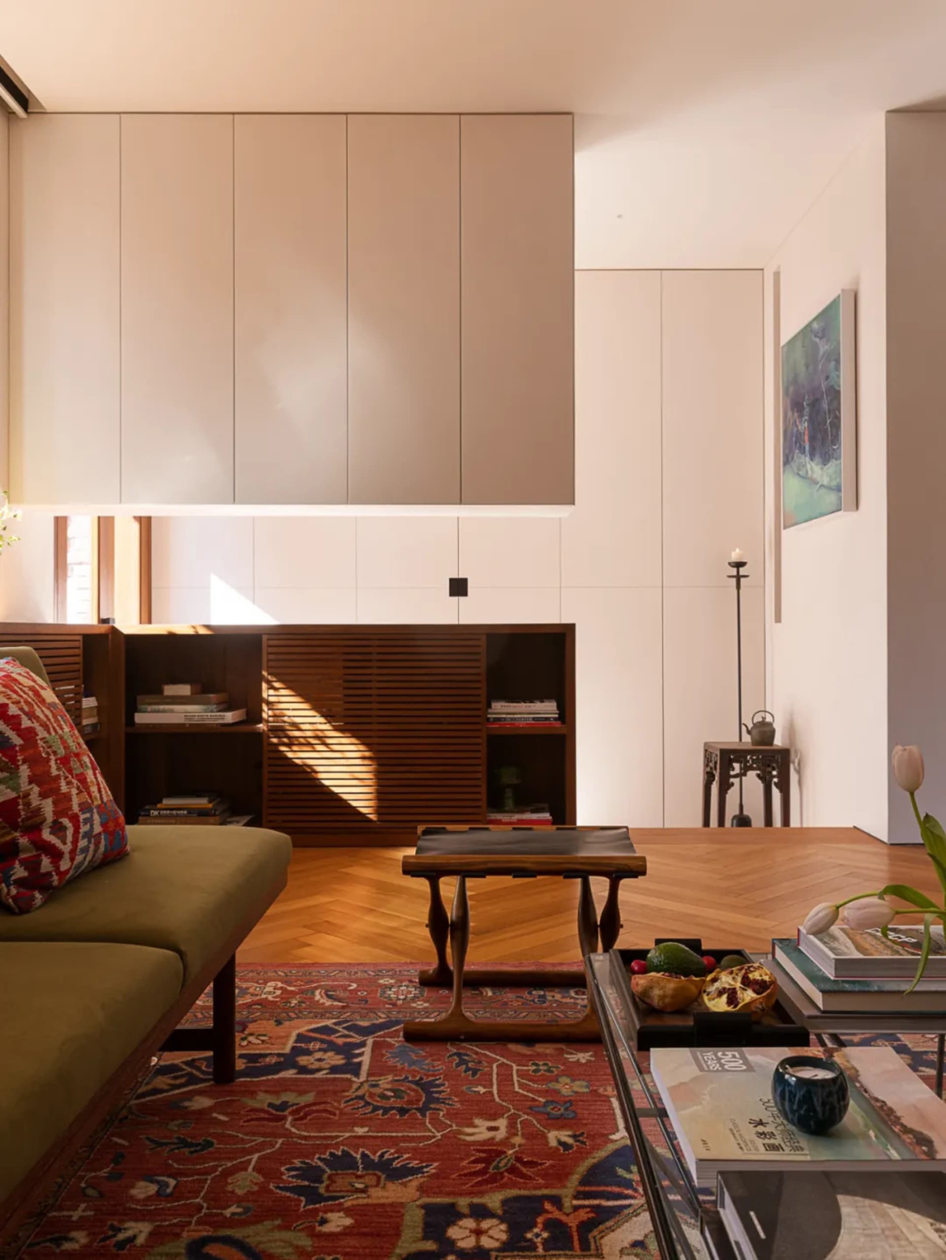 Modern minimalist living room featuring built-in beige upper cabinets, walnut lower storage with louvered doors, olive green sofa with patterned cushion, Persian rug on herringbone wood floor, and natural sunlight streaming in.