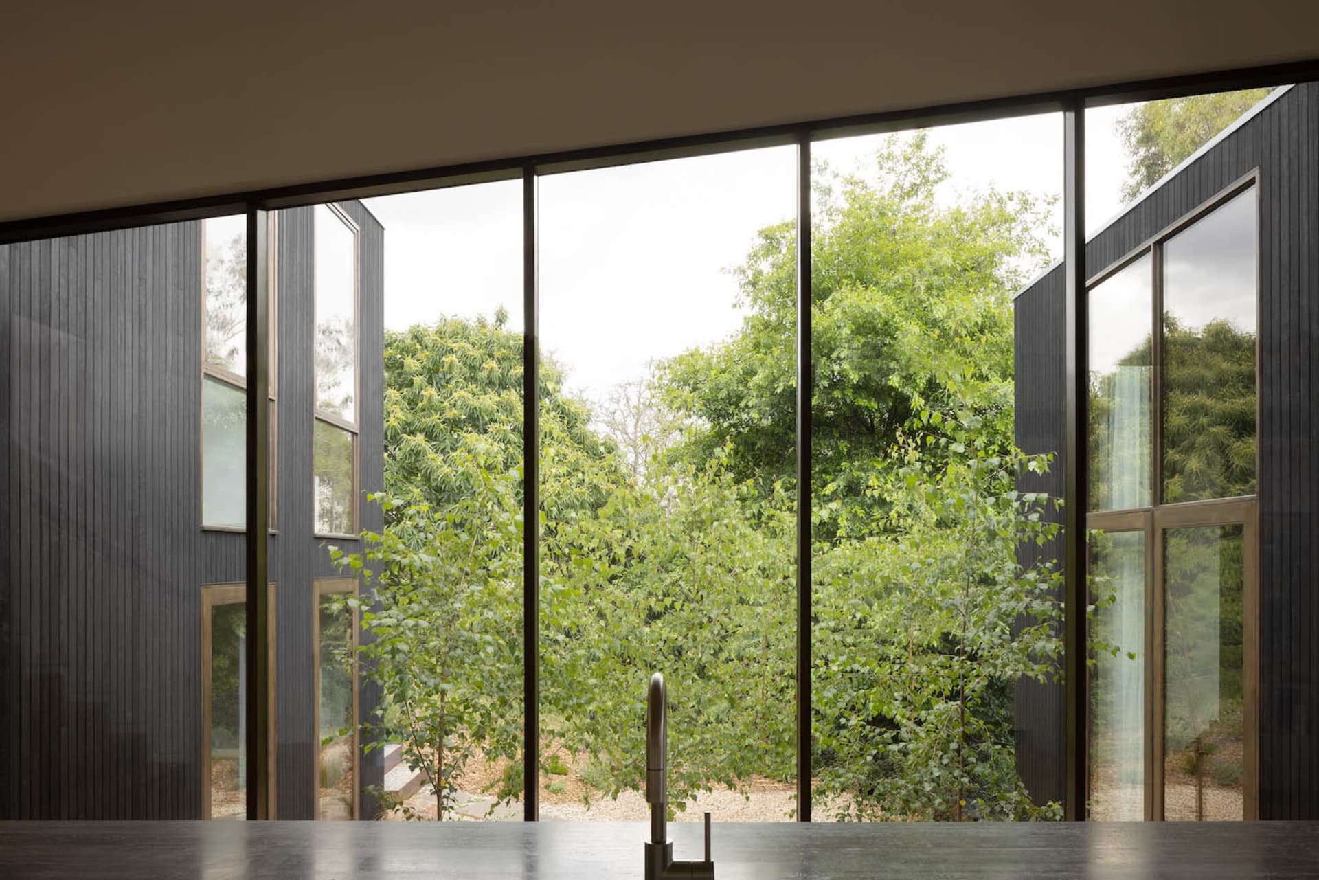 Modern kitchen interior featuring a dark countertop, matte black faucet, and large panoramic windows overlooking a dense green forest with dark vertical wood cladding on the exterior walls.