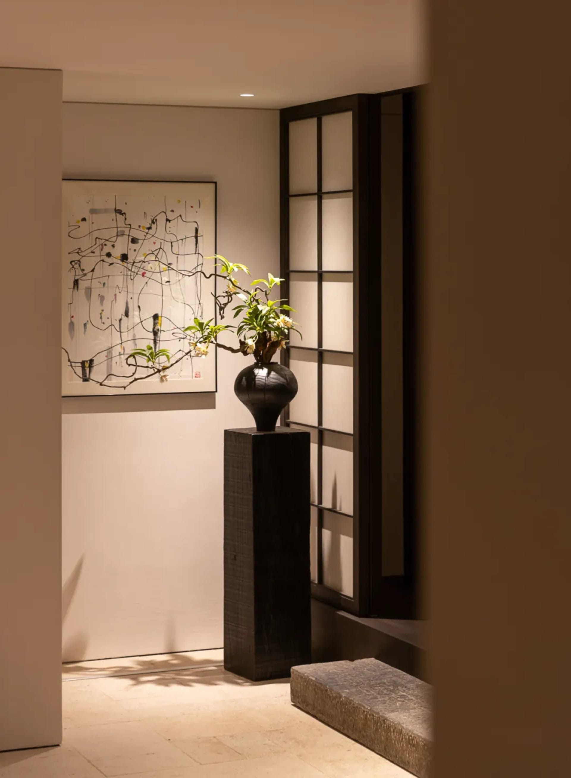 現代極簡玄關,深色木座台上擺放花道插花作品,牆上掛有抽象水墨畫,旁邊是黑色日式格柵屏風,暖光照明。
