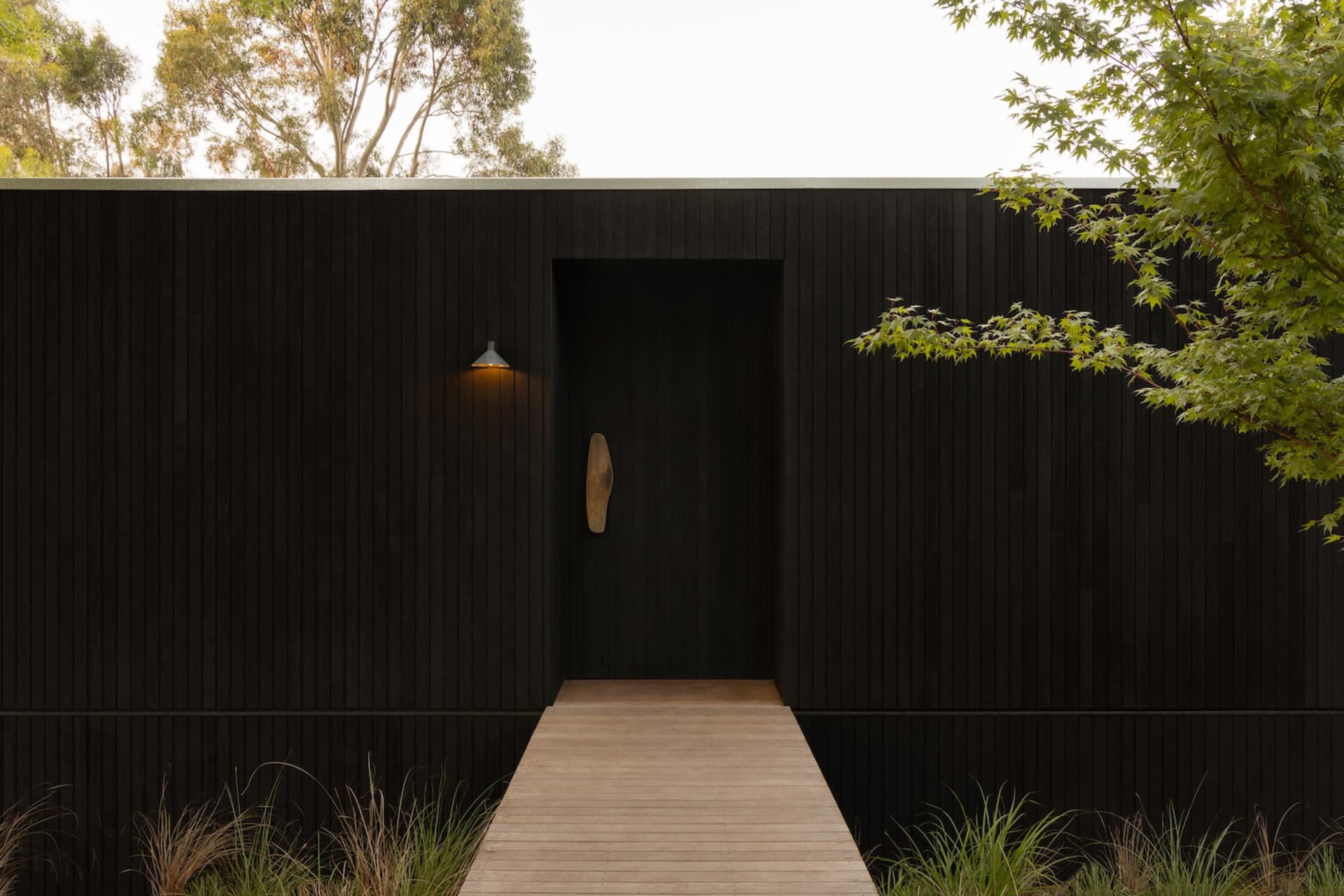 Front entrance of a modern house with black charred wood cladding, a wooden boardwalk path, wall sconce lighting, and surrounding greenery.