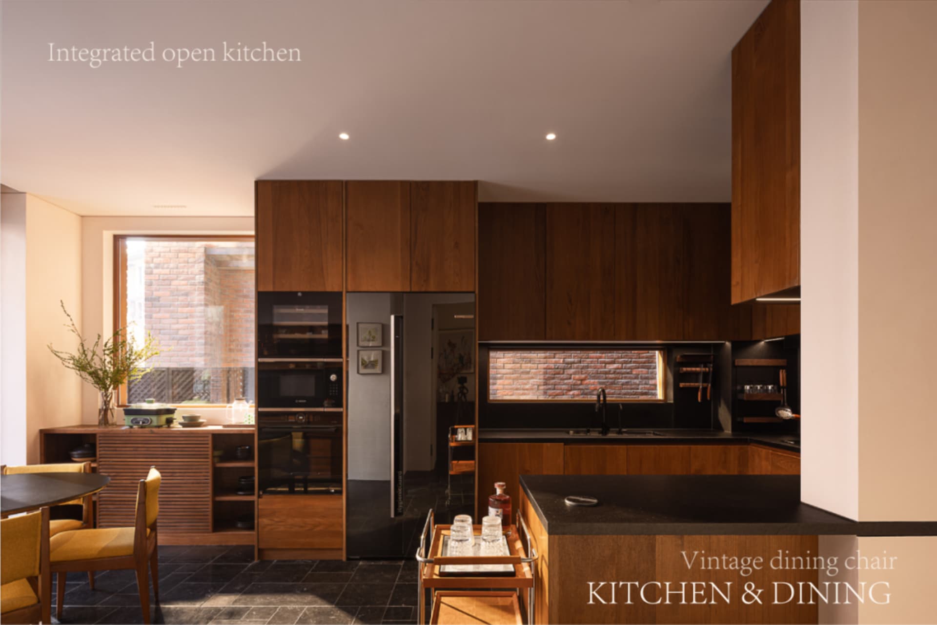 Modern open-plan kitchen featuring rich walnut wood cabinetry, black stone countertops, built-in appliances, and a vintage-style dining area with mustard-yellow upholstered chairs.