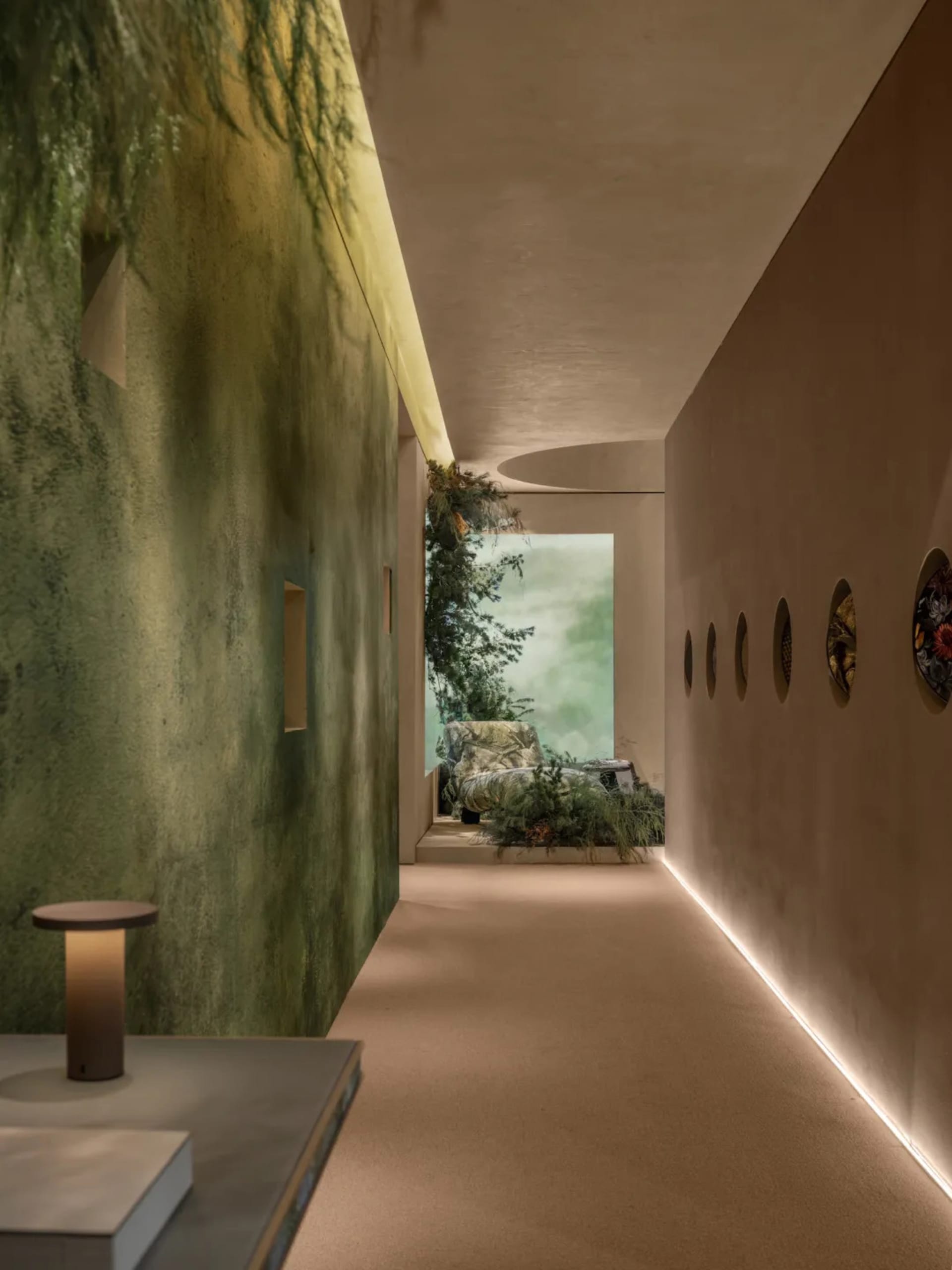 Modern interior hallway featuring a textured green wall, integrated greenery, circular wall art, and a distant reading nook with natural light.