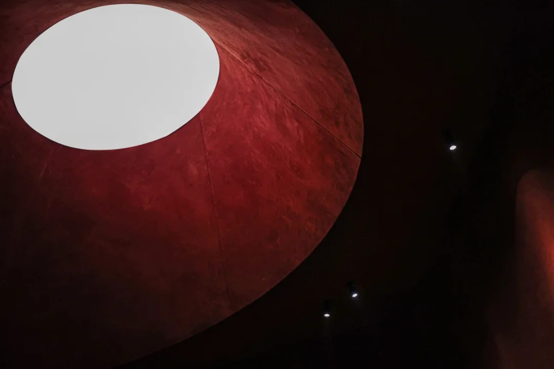 Low-angle view of a large, curved ceiling finished in textured deep red plaster with a prominent circular white oculus emitting bright light, accompanied by small spotlights in the dark background.