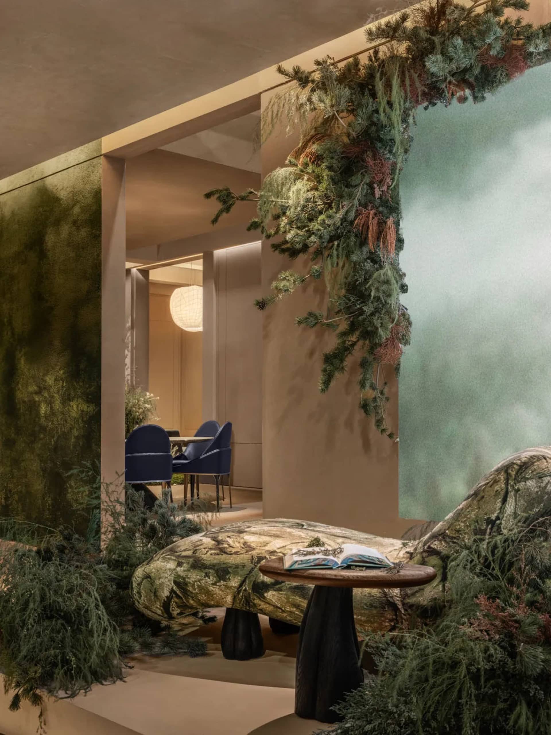 Modern biophilic lounge featuring a moss-textured chaise lounge, dark wood side table with open book, abundant hanging coniferous greenery, textured green wall, and a dining area visible in the background with blue velvet chairs.
