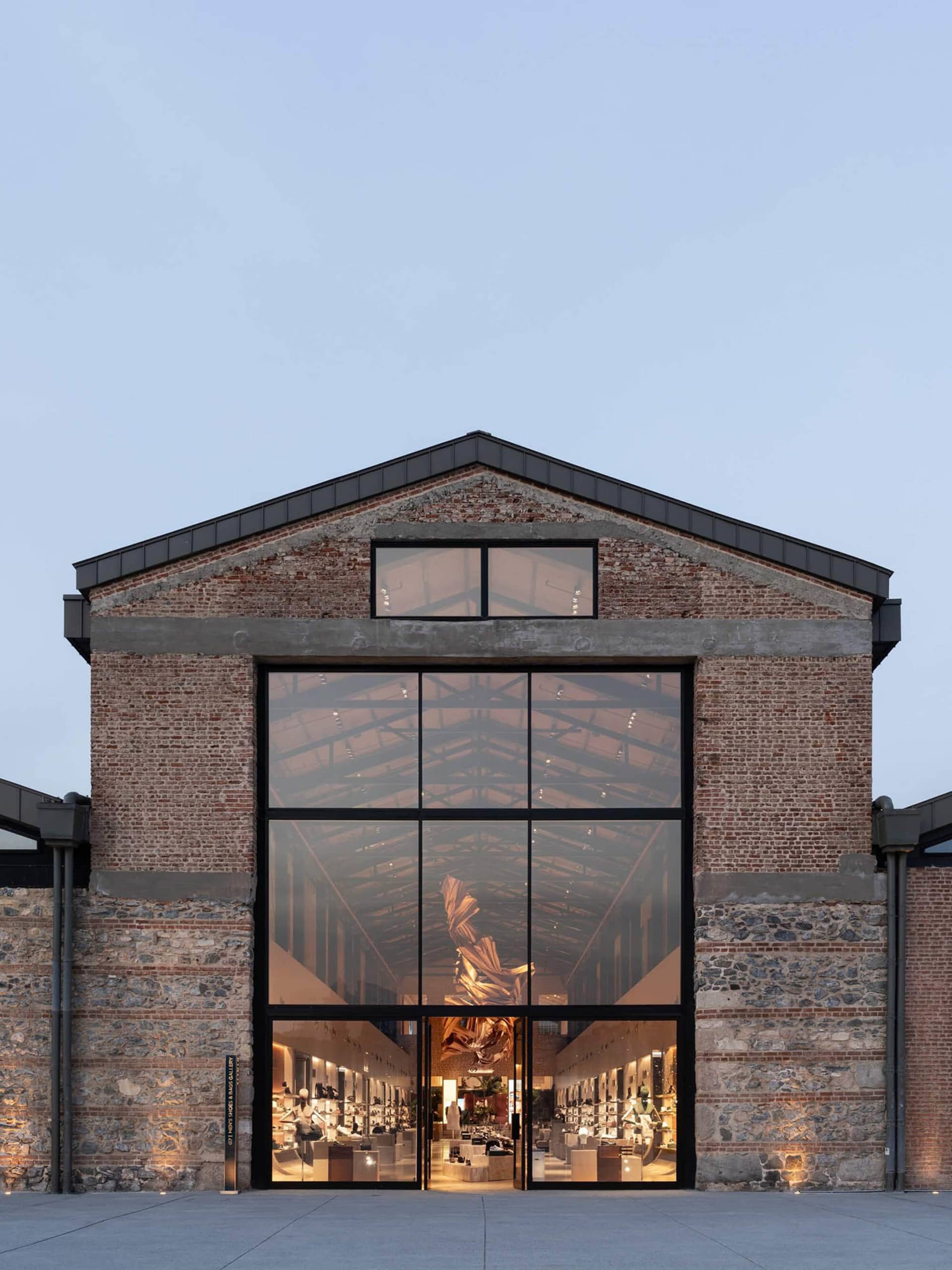 Front exterior view of a luxury retail store with a large black-framed glass facade set within a restored red brick and stone industrial building at dusk.