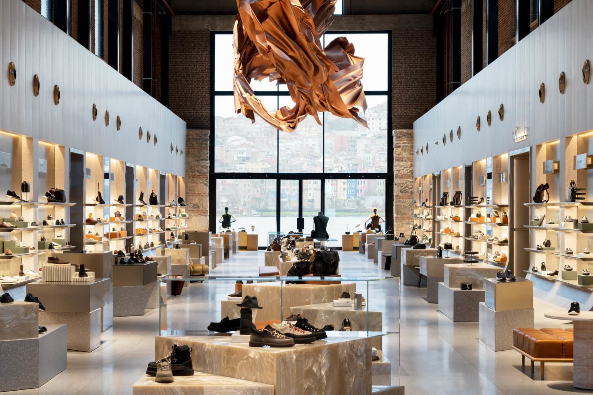 Wide view of a modern luxury shoe and handbag store featuring white walls, exposed brick, terrazzo display platforms, and a large abstract copper sculpture hanging from the ceiling with a cityscape visible through tall windows.