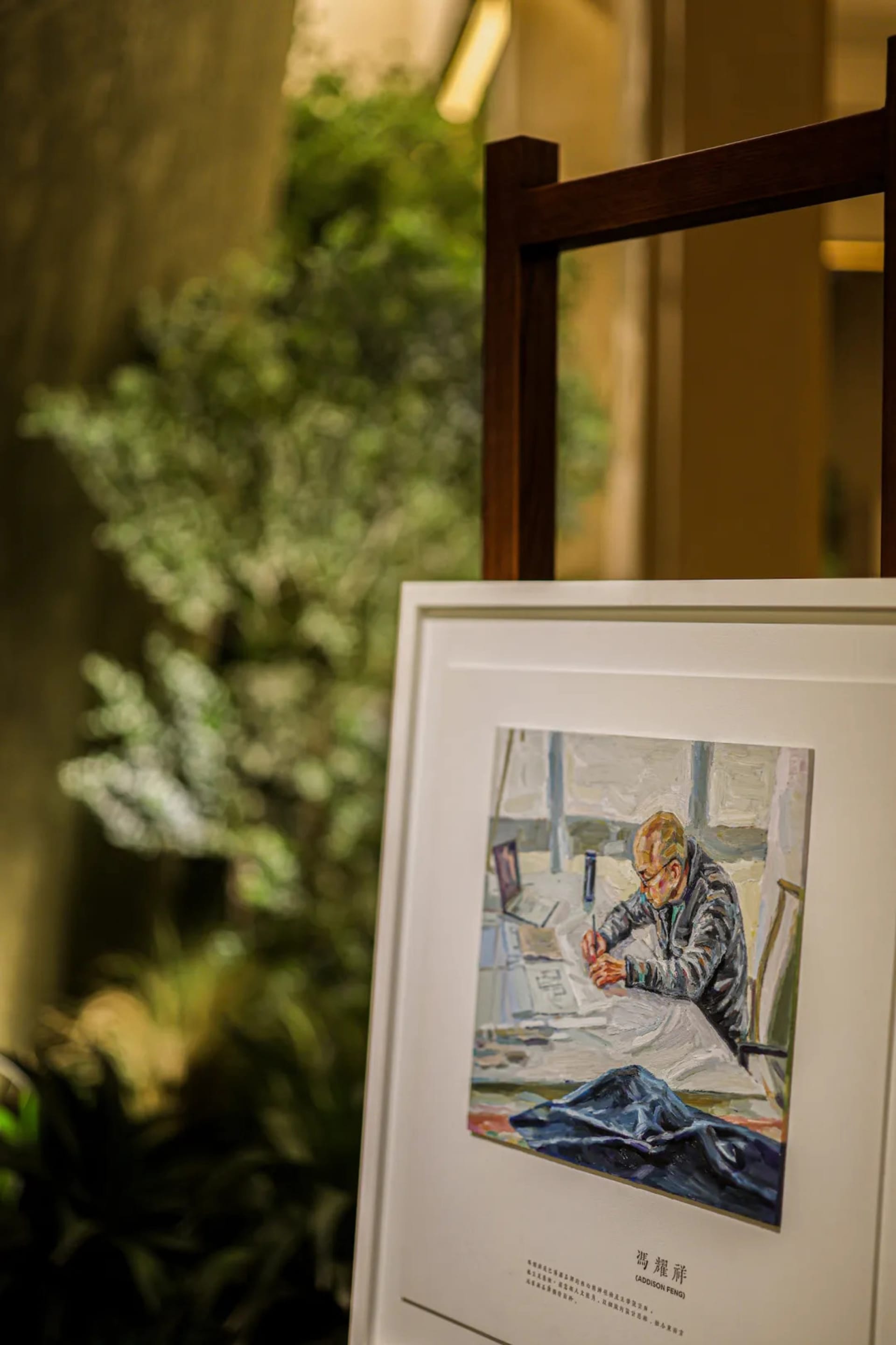 A framed oil painting depicting an older male artist working at his desk is displayed on a dark wooden easel in a modern interior with blurred green foliage in the background.