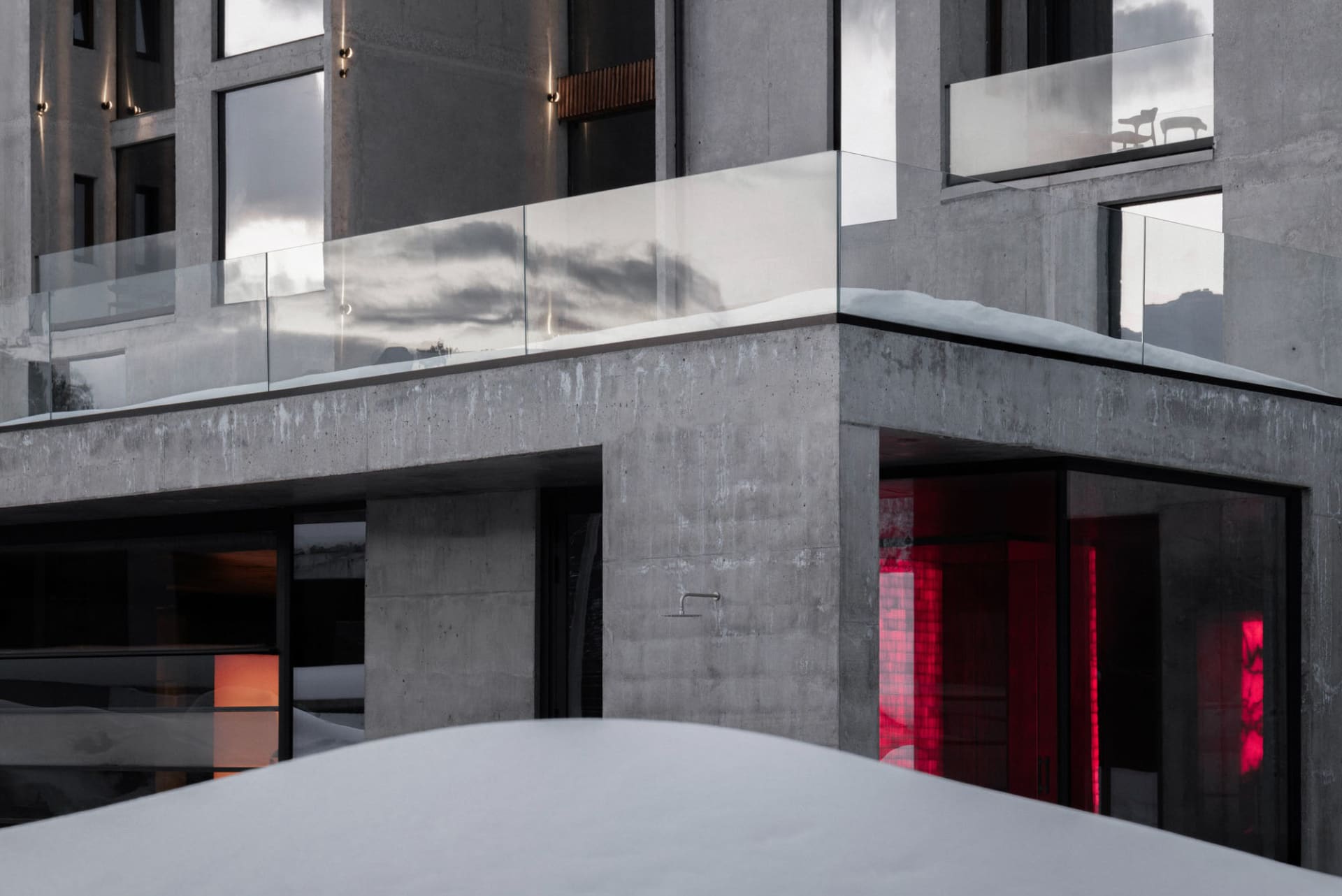 Exterior view of a modern brutalist concrete house featuring large glass windows, a snow-covered cantilevered glass terrace, and dramatic red interior lighting visible through lower-level openings.