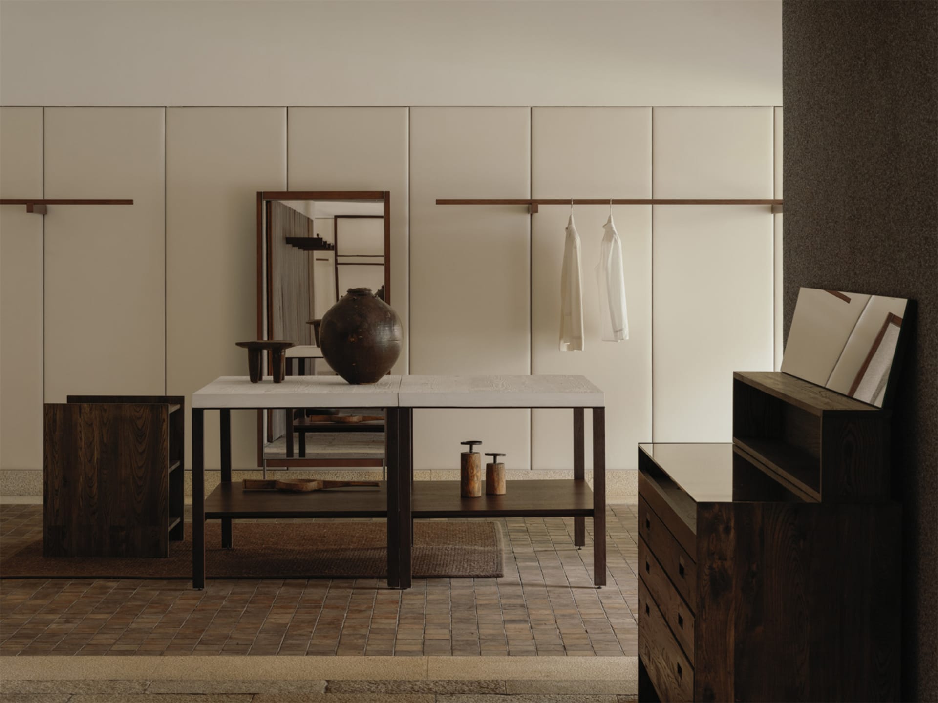 Modern minimalist entryway with dark walnut console table, large ceramic vase, wooden mirror, and hanging white garments against beige paneled walls.