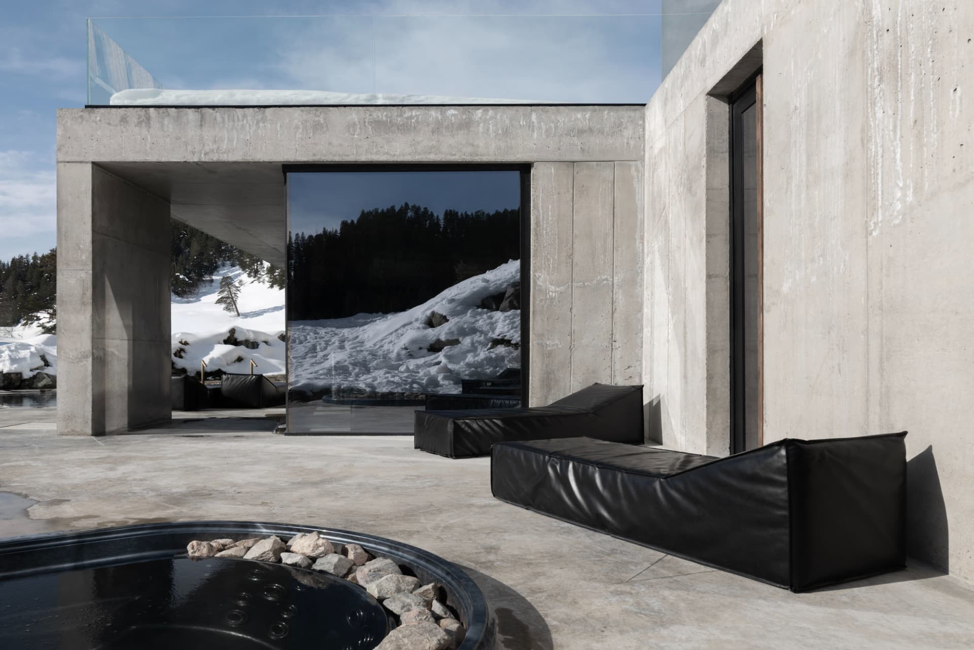 Modern concrete villa exterior with black leather daybeds, circular hot tub, and large glass windows overlooking snowy mountain landscape.