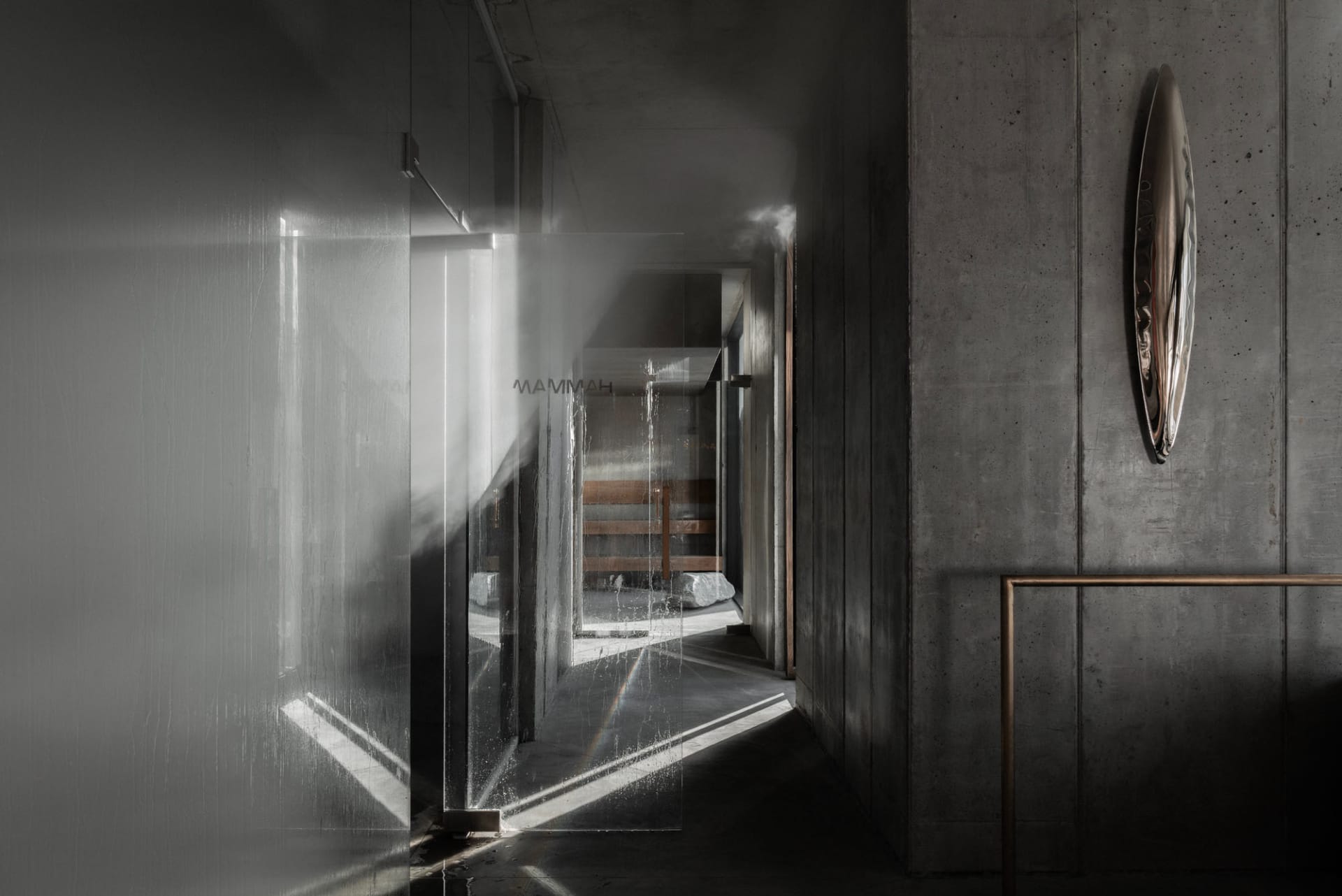 Narrow brutalist concrete hallway featuring a large translucent glass partition wall, dramatic diagonal light beams, and an asymmetrical metallic wall sculpture.