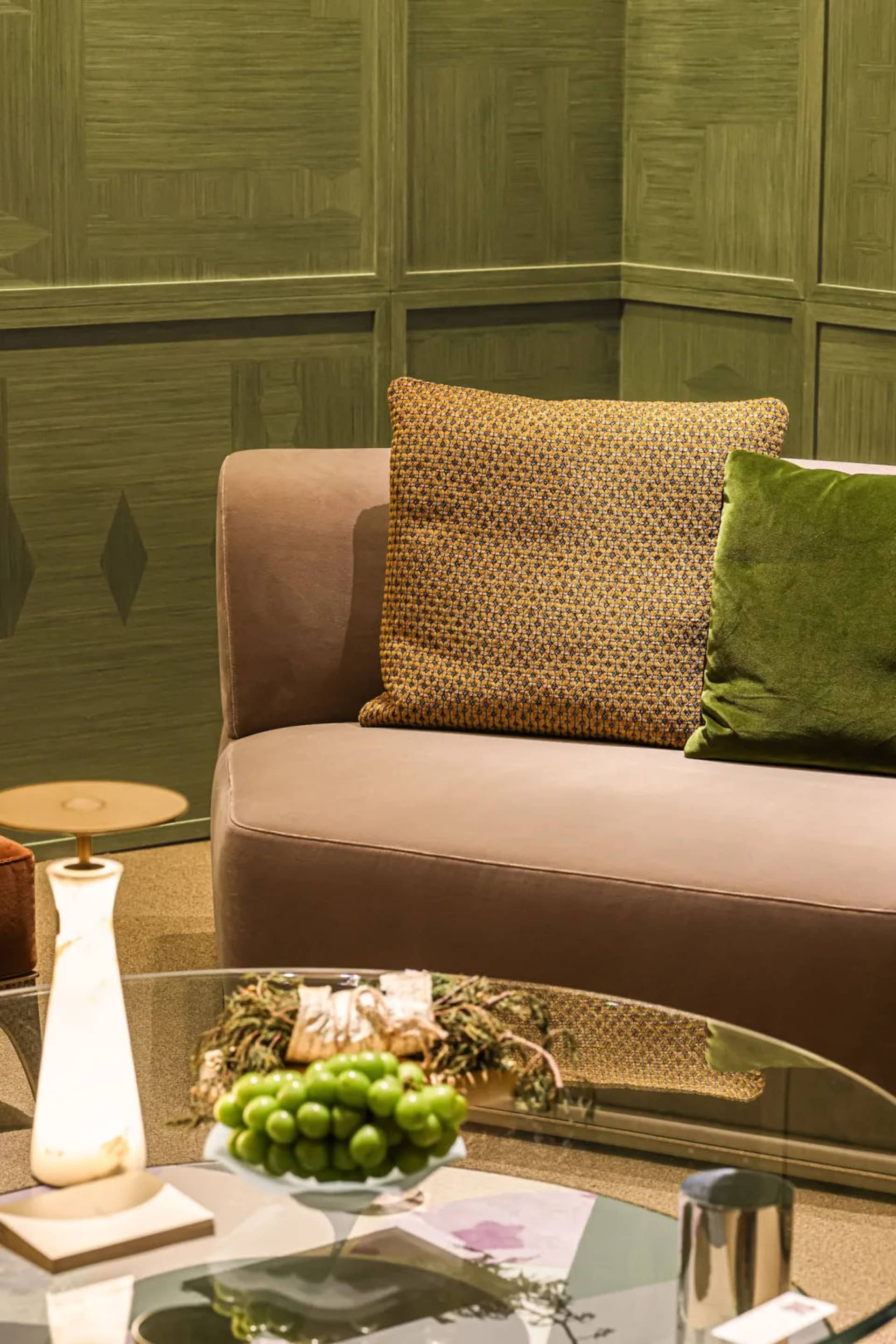 Close-up of a modern living room corner with a taupe velvet sofa, mustard patterned pillow, green velvet pillow, and an olive green textured wall panel background. A glass coffee table in the foreground holds a bowl of green grapes and a marble lamp.