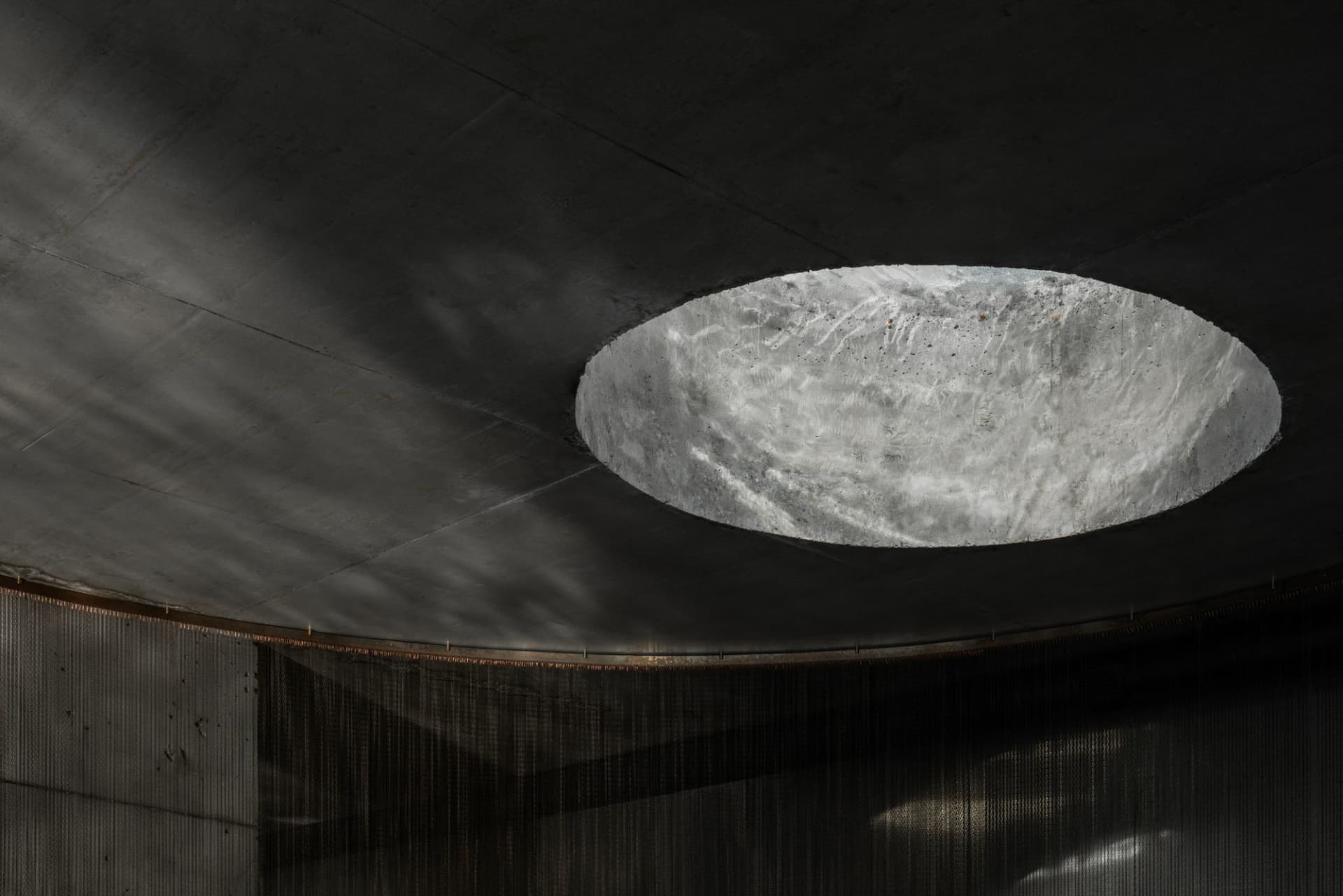 View looking up at a large circular concrete oculus skylight set into a dark raw concrete ceiling with hanging chainmail curtain below.