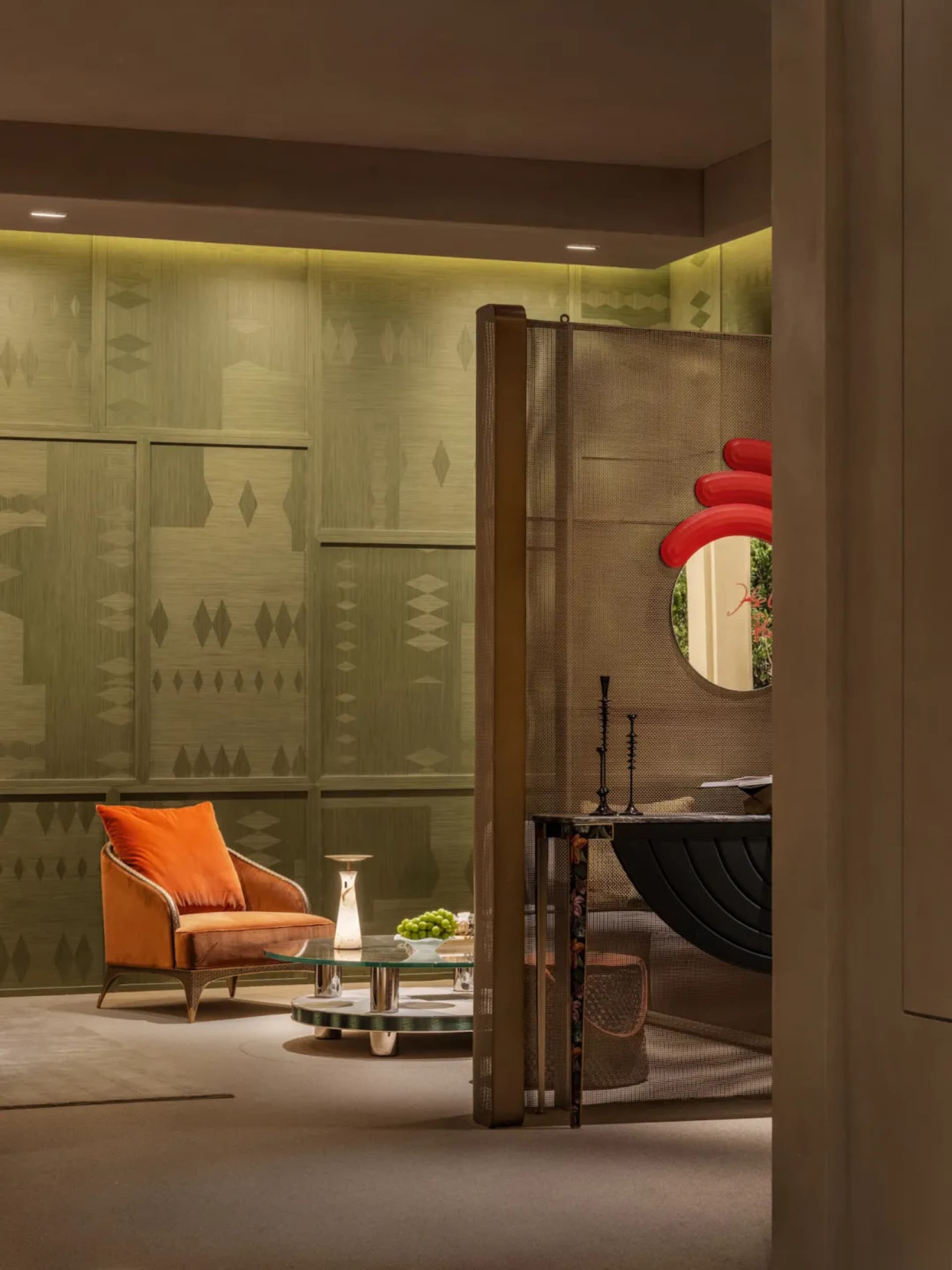 Modern living room featuring sage green patterned wall panels, burnt orange velvet armchair, round glass coffee table, and a bronze mesh room divider with red circular mirror.