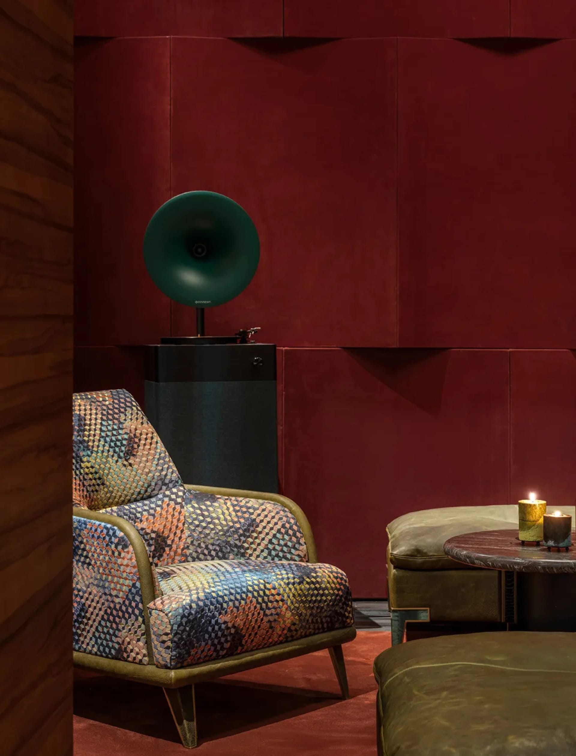 Colorful geometric patterned armchair with olive green frame placed against deep red padded wall panels, accompanied by a vintage green gramophone on a black pedestal and leather ottomans with lit candles.