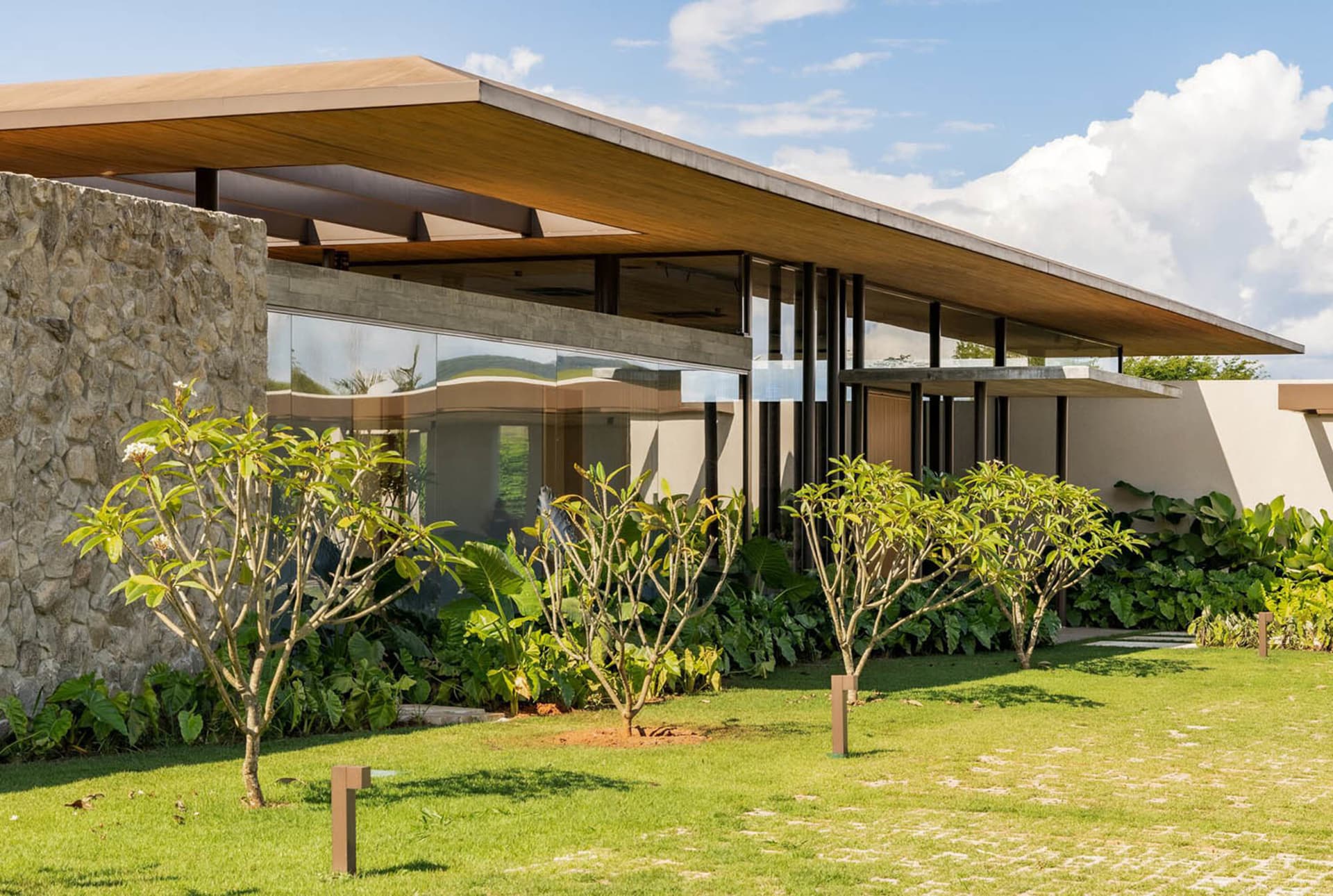 Modern tropical house exterior featuring a large stone wall, extensive wood overhang, floor-to-ceiling glass windows, and tropical landscaping with plumeria trees on a sunny day.