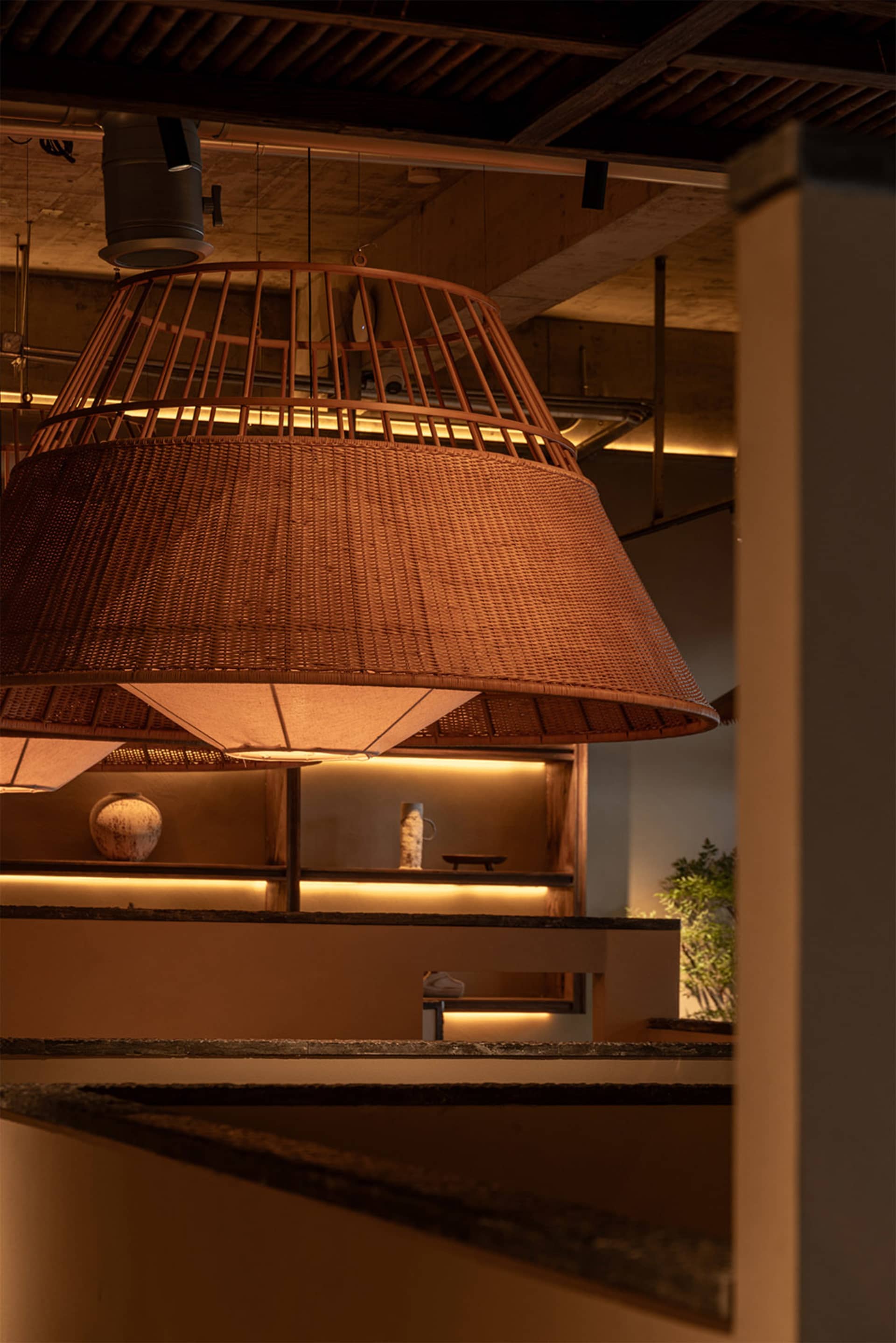 Large conical woven bamboo pendant lamp with warm internal lighting hanging above a dark stone counter in a modern Japanese-inspired interior with concrete ceiling and backlit wooden shelves.