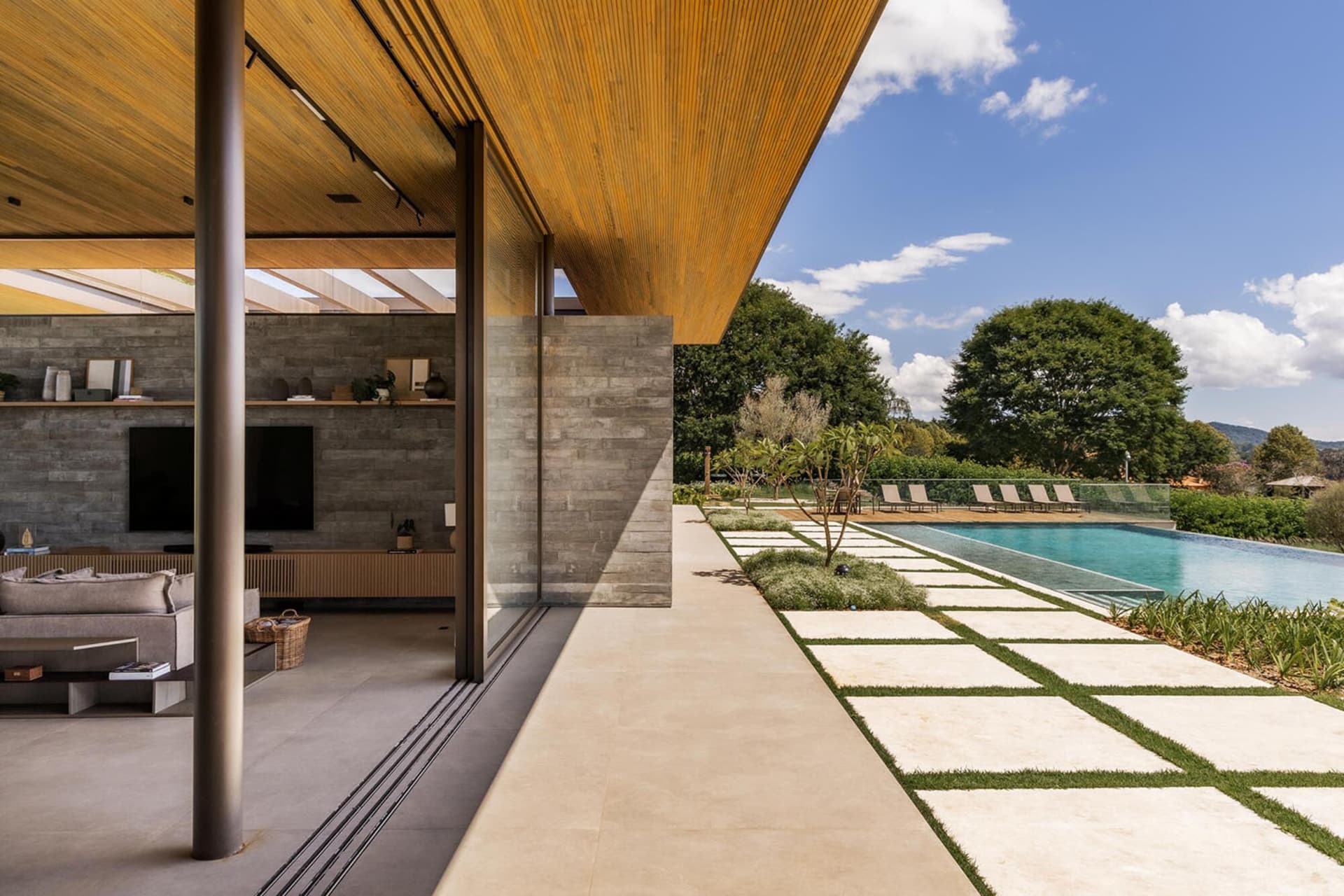 Modern villa covered terrace featuring a warm wood slat ceiling, textured stone wall, and an infinity pool with stone paver walkway extending into a lush garden under a blue sky.