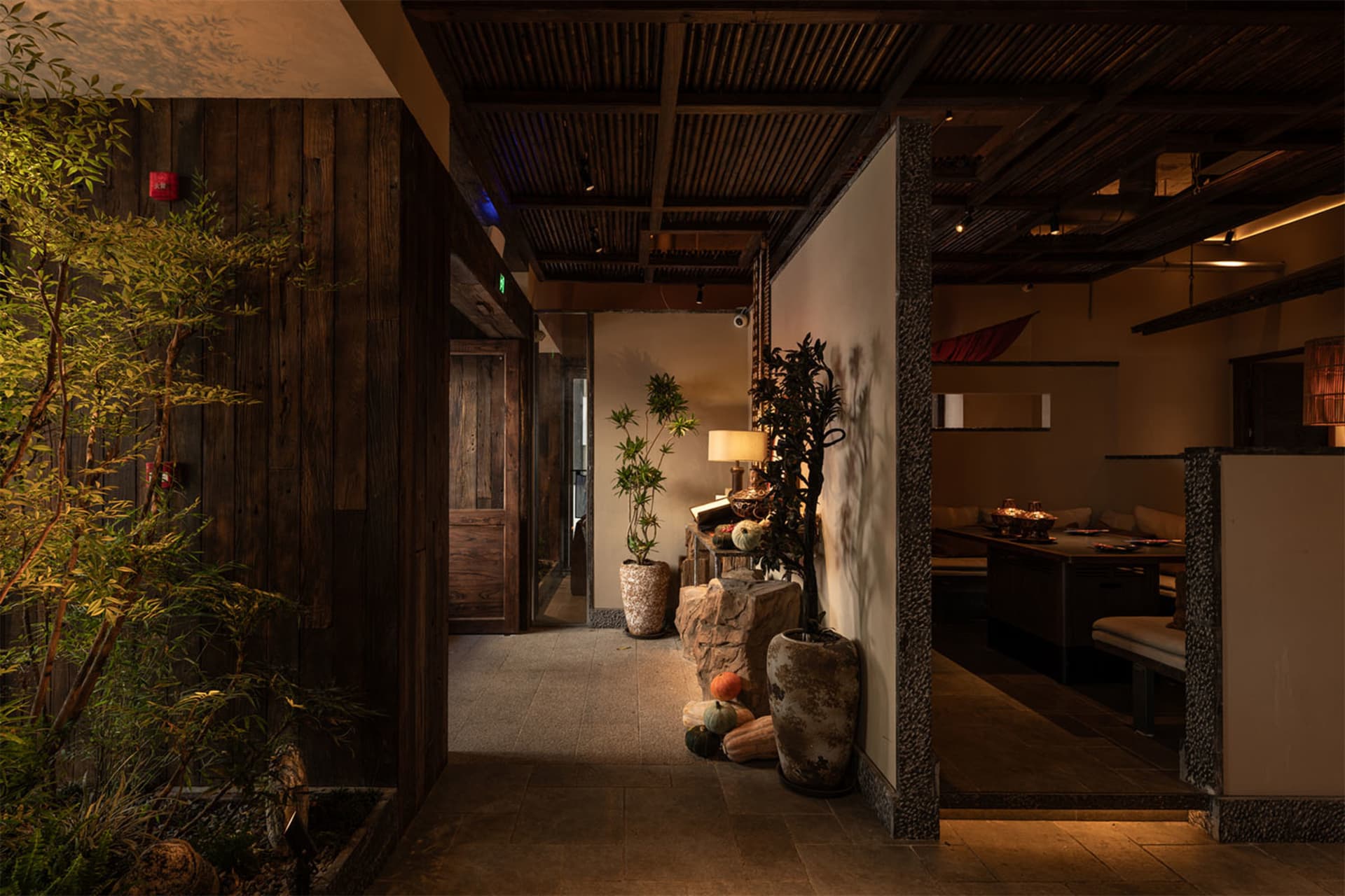 Dimly lit hallway in a Japanese restaurant featuring dark wood walls, textured stone pillars, potted plants, and a view into a warm dining area with banquette seating.