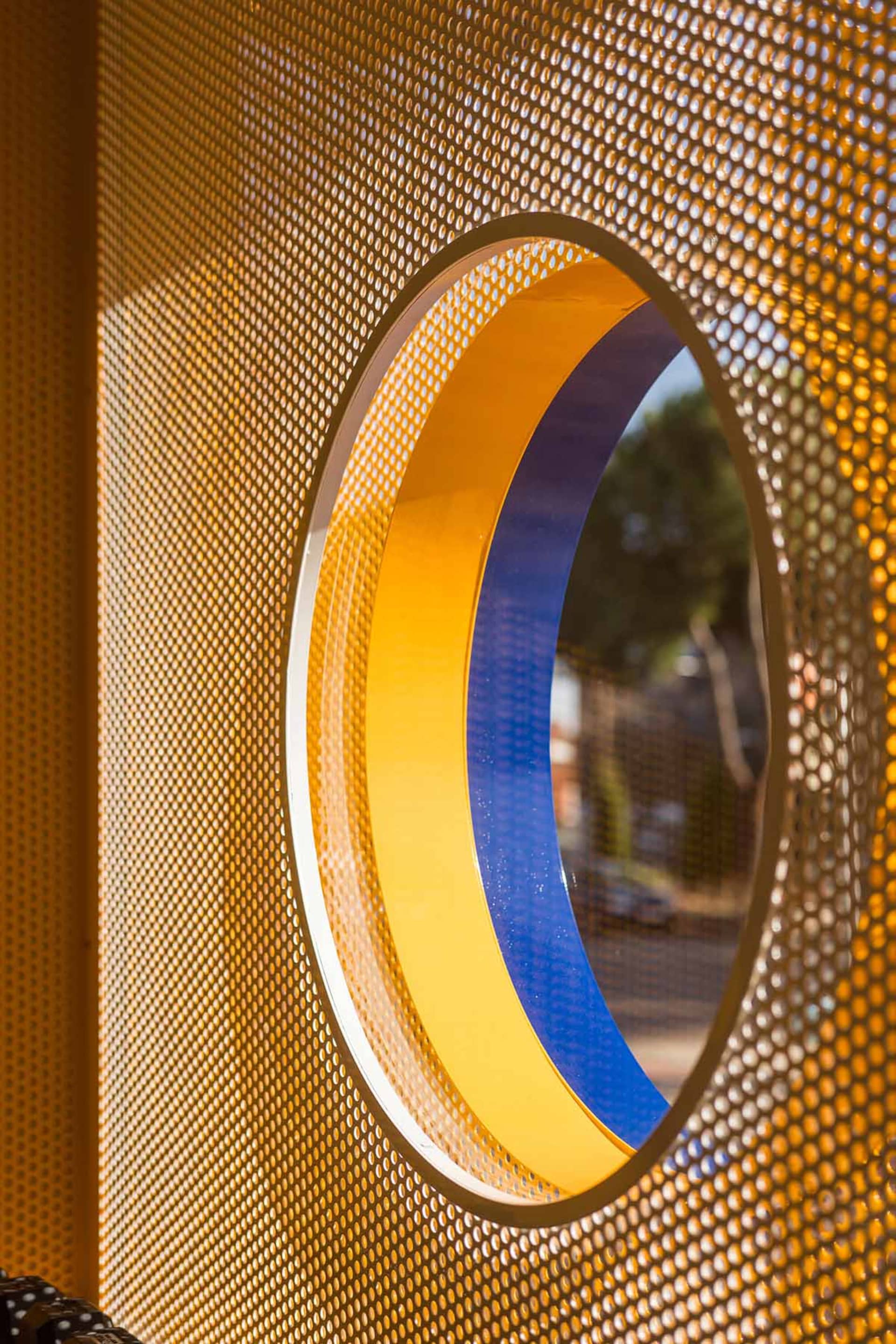 Close-up of a circular opening with yellow and blue painted rings set within a golden perforated metal screen, with trees visible in the background.