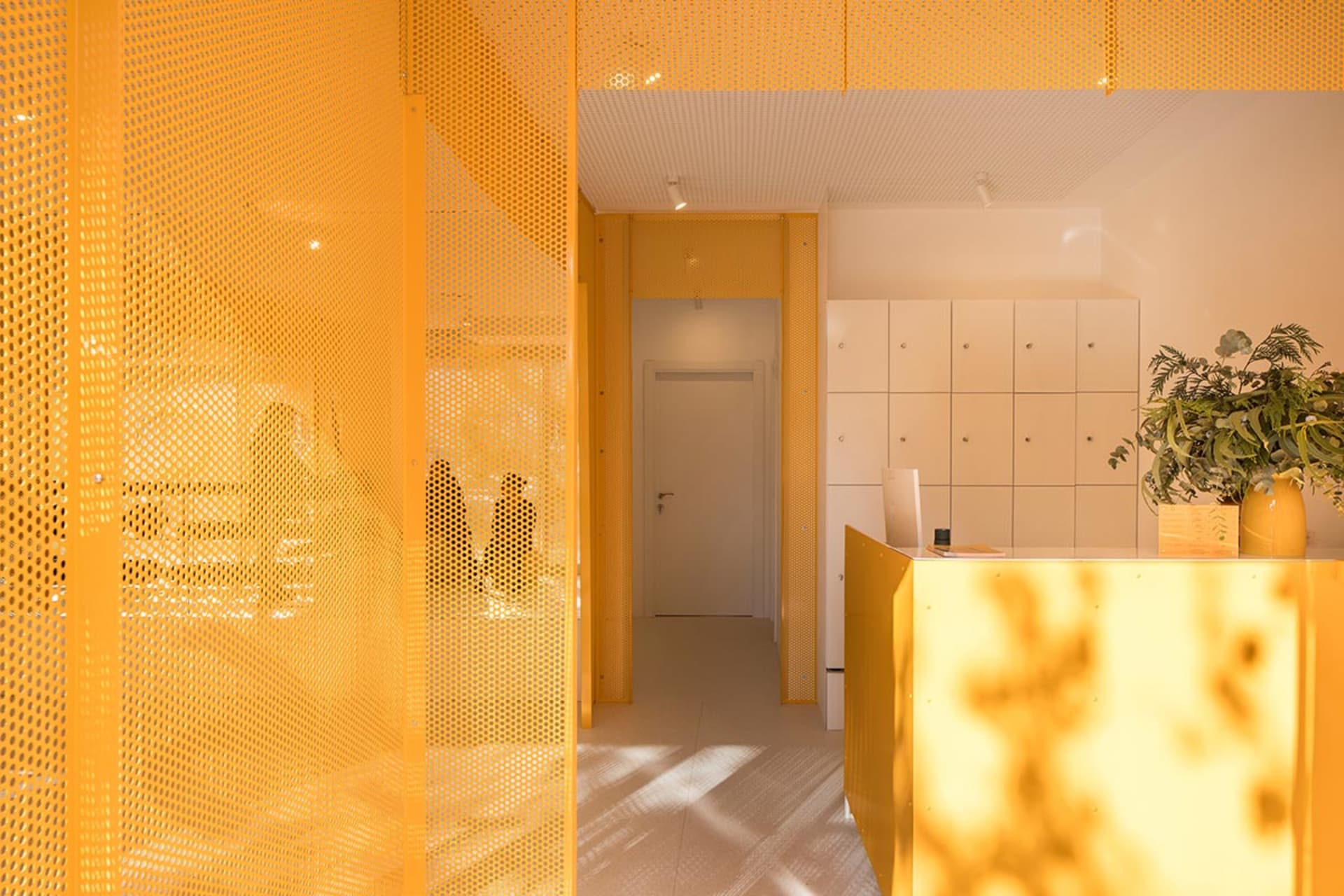 Modern reception area featuring bright yellow perforated metal screens, a matching yellow counter, white lockers, and a potted plant in a yellow vase.