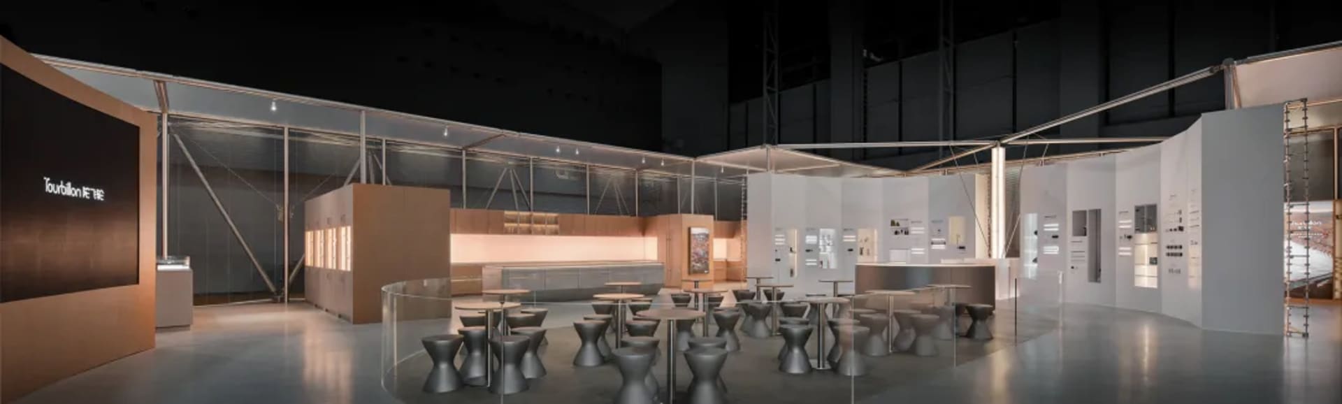 Modern industrial-style exhibition hall with a cafe area containing numerous grey concrete stools and tables arranged in an open layout, surrounded by display walls and glass partitions.