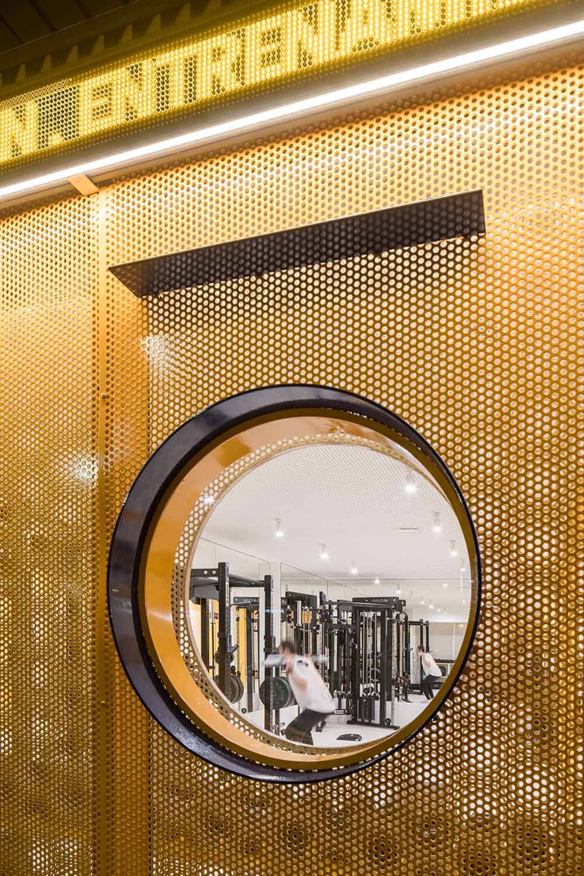 Circular mirror set in a golden perforated metal wall overlooking a modern gym with people lifting weights.