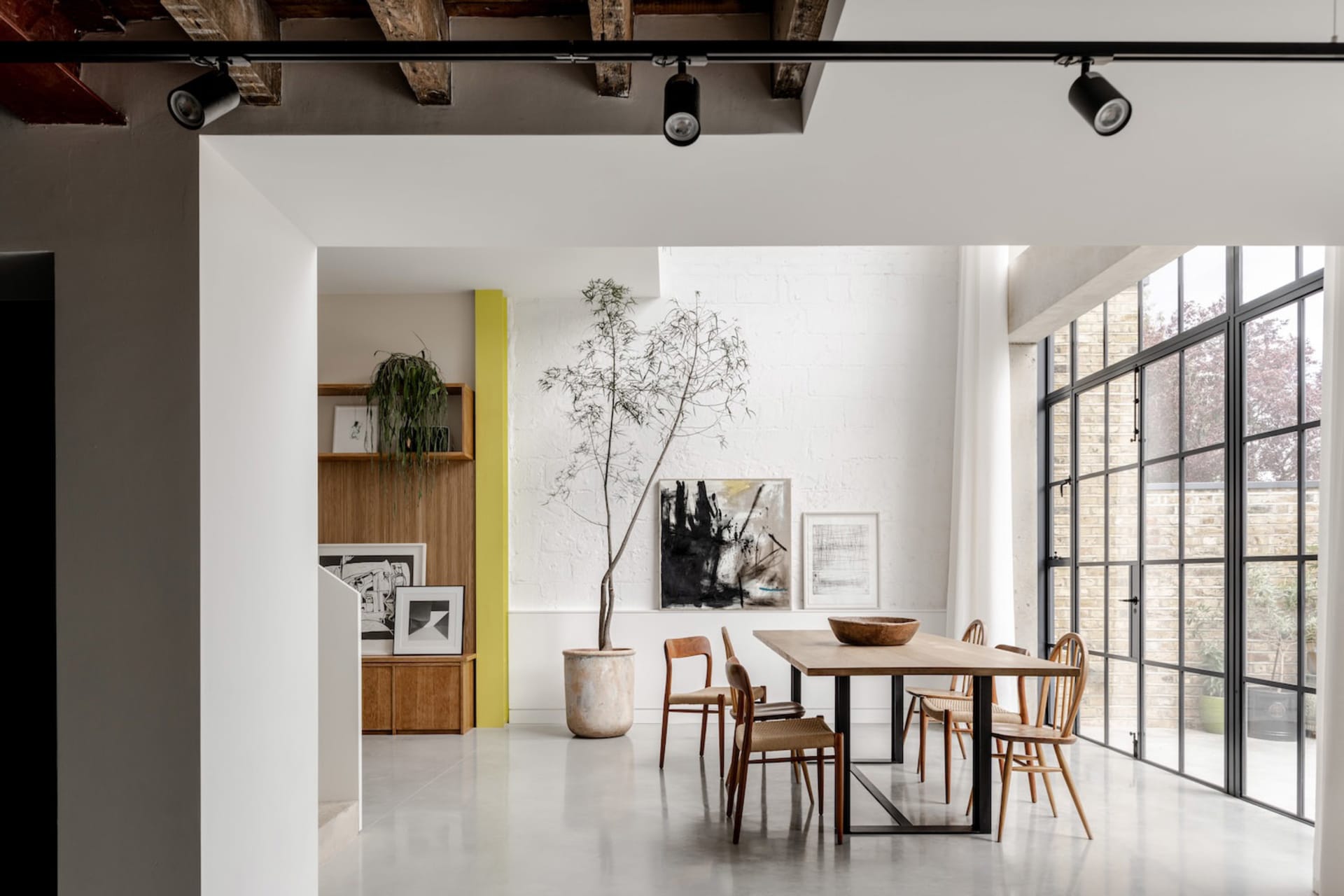 Modern industrial loft dining room featuring a long oak dining table with mixed wooden chairs, a large potted tree, white brick walls, and large black-framed windows allowing abundant natural light.