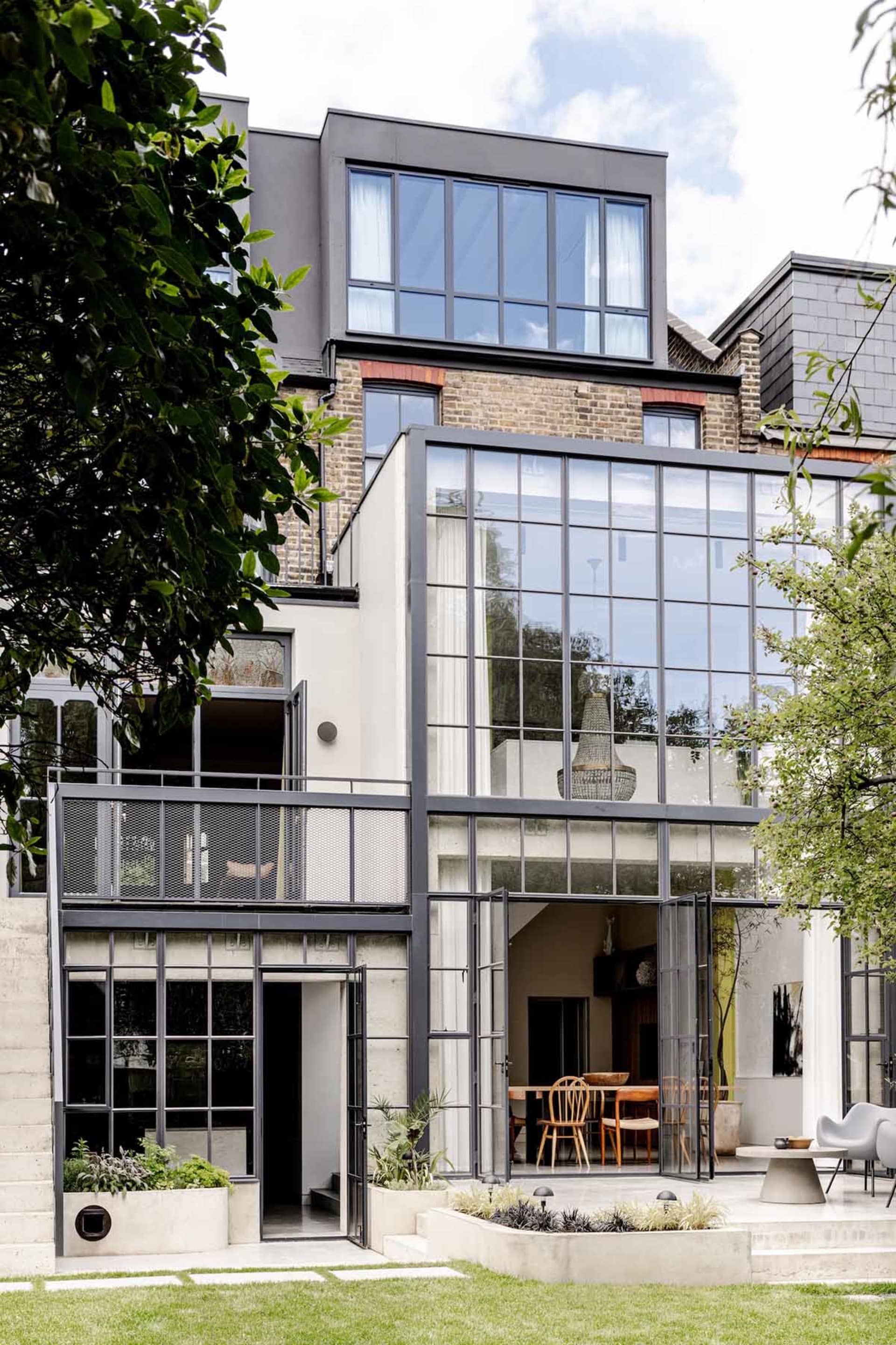 Modern two-storey glass and steel extension of a period house featuring large pivot doors opening to a landscaped garden with concrete planters and outdoor dining area.