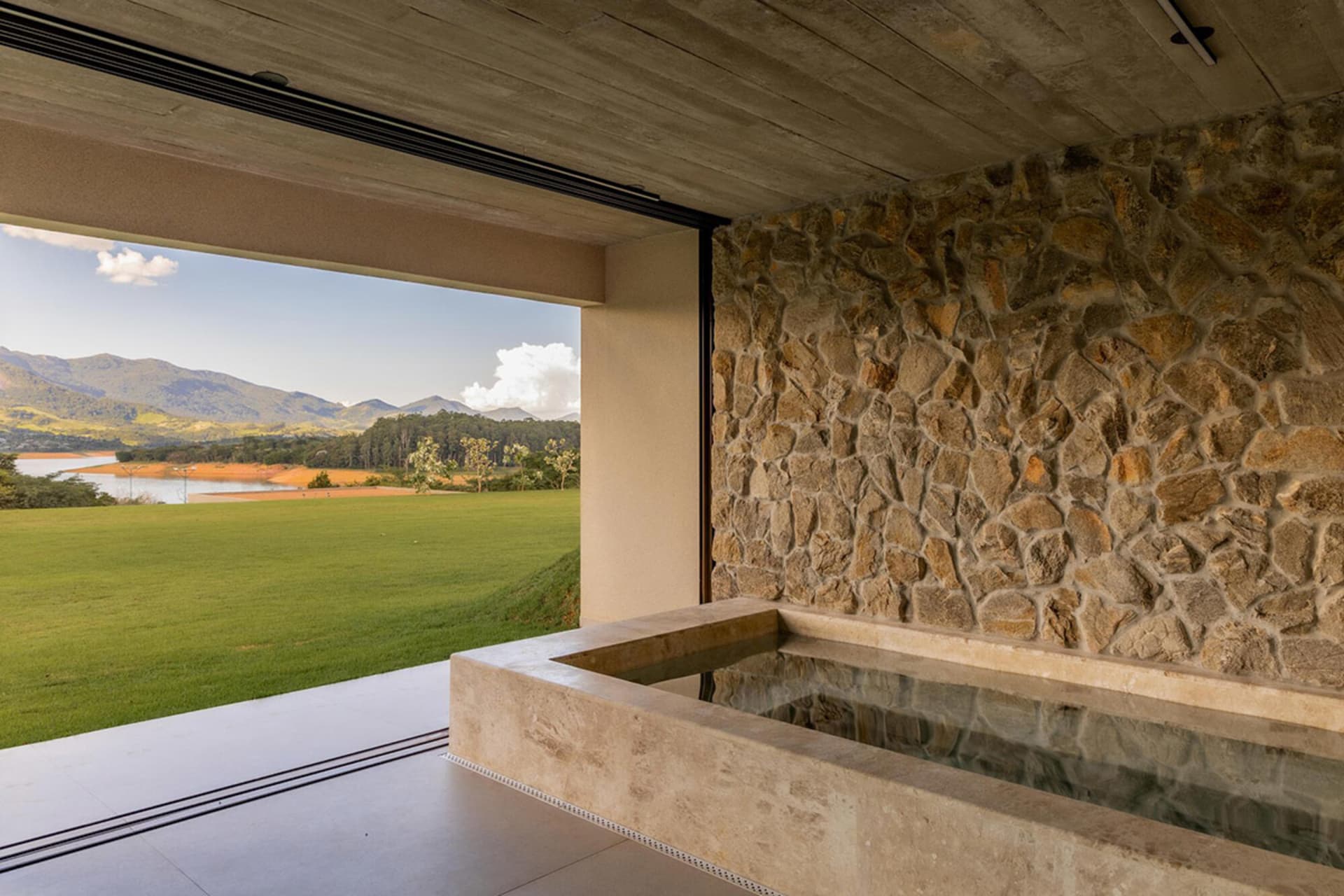 Modern interior with a long stone water feature against a rough natural stone wall, opening to a wide green lawn, lake and mountain range under a blue sky.