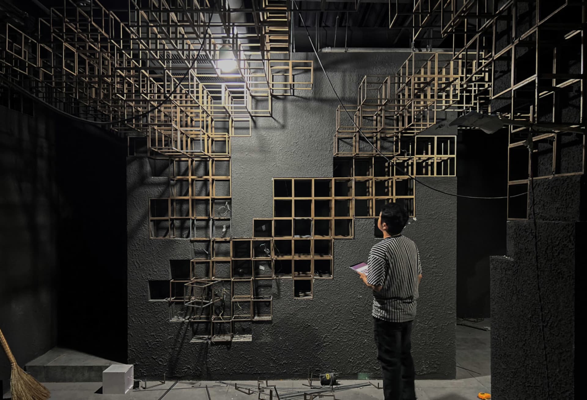 Man in striped shirt observing a large-scale wooden cube grid installation mounted on a dark concrete wall in an industrial exhibition space with dramatic overhead lighting.