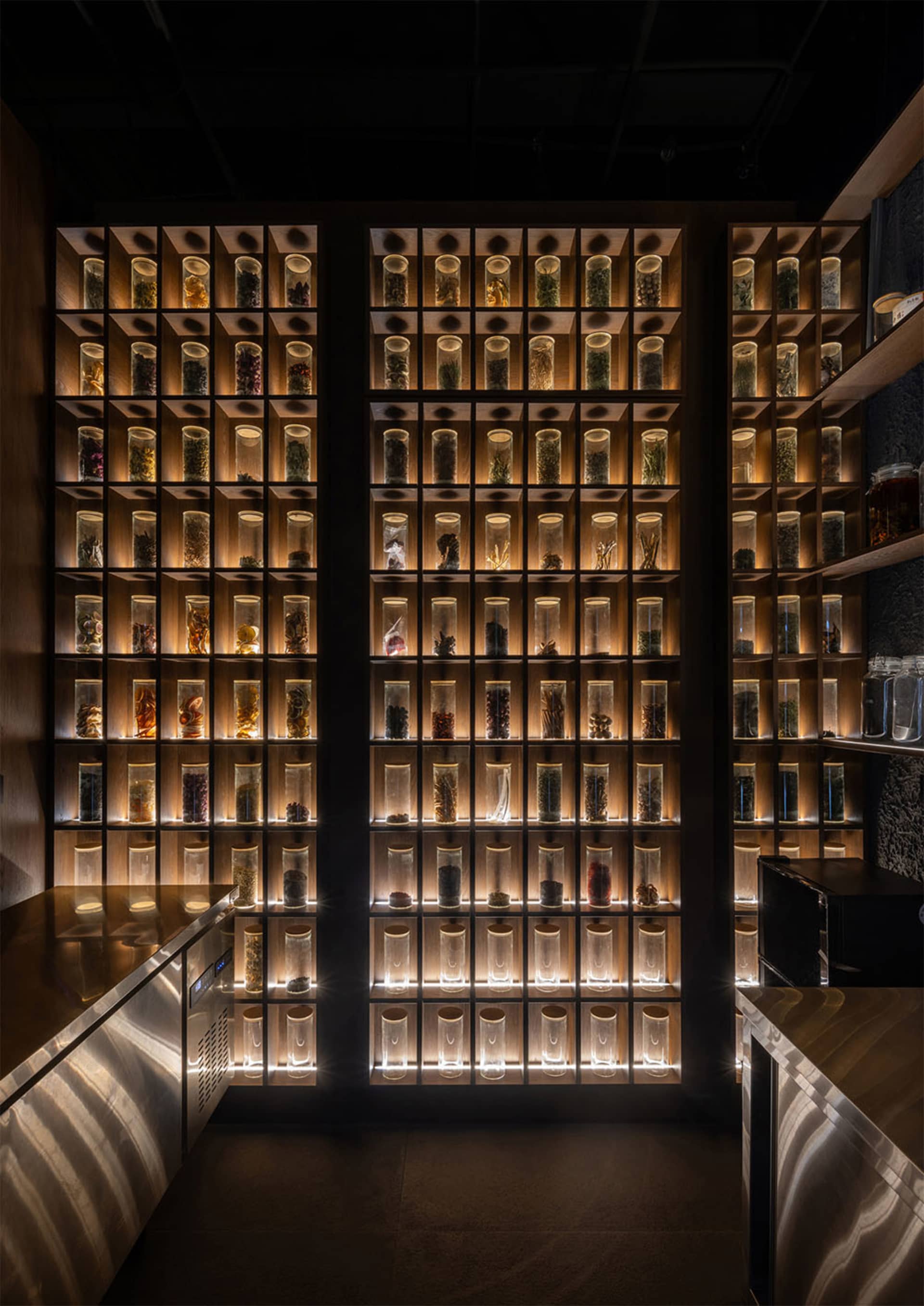 Modern apothecary wall with backlit walnut wood cubbies filled with glass jars of dried herbs and botanicals in a luxury tea or wellness space.